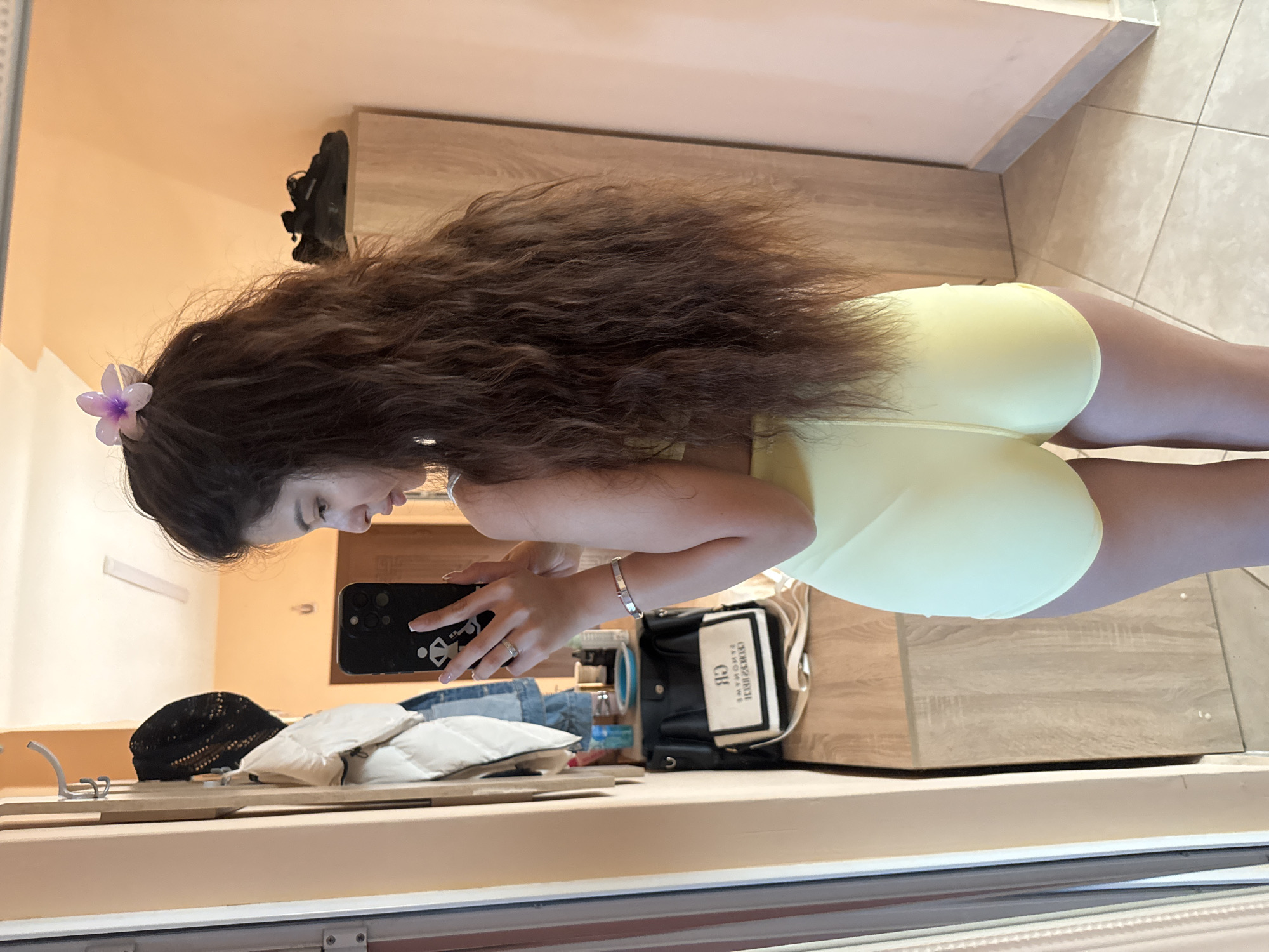 A woman with long, wavy brown hair, adorned with a small purple flower, is taking a photo of herself with a black smartphone in what appears to be a hotel room or similar accommodation. She is wearing a light yellow outfit that seems to be exercise or swimwear. In the background, there's a shelf with a black and white purse, a puffy white coat or blanket, a black hat, and a pair of blue jeans. The setting includes a light-colored wooden floor, wardrobe and wall painted in light orange. The location appears to be indoors, in a room. The specific location within the city of Plovdiv, Bulgaria is not discernible from the image.