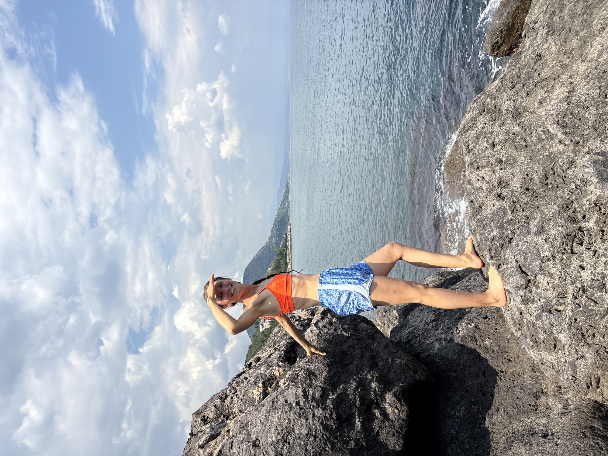 A young woman stands on dark, rugged rocks by the sea under a cloudy sky. She is wearing an orange bikini top and blue and white patterned shorts. Her right hand is raised to shield her eyes as she looks out at the water. Her left arm is extended back, with her hand resting on a large rock to her left. The sea is a deep blue, with gentle waves lapping at the shore. In the distance, a green, hilly coastline is visible, with some buildings nestled on the slopes. The sky is filled with a mix of white and gray clouds, with patches of blue sky showing through. The scene is set in Bondarenkovo, Ukraine.