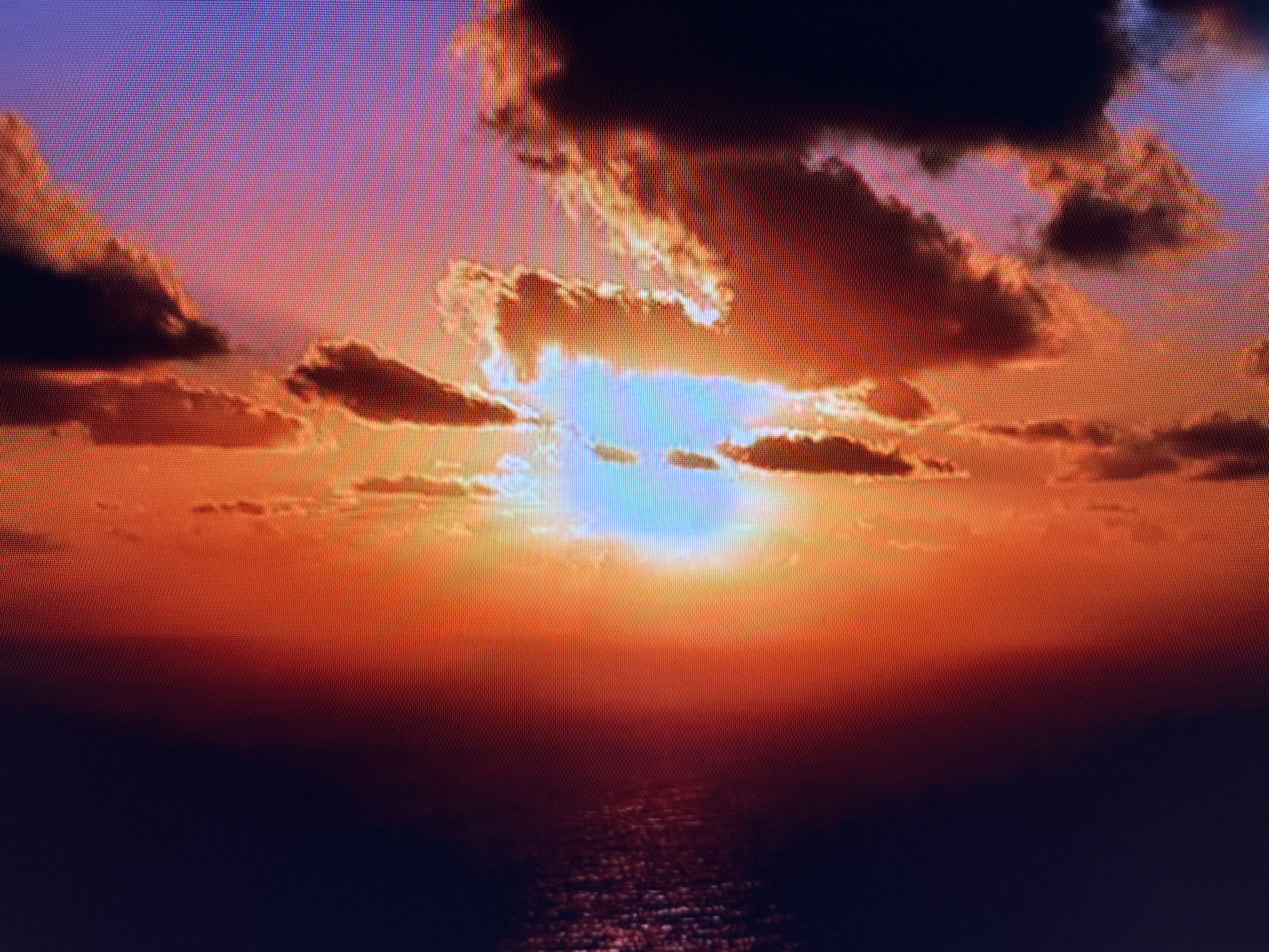 The image depicts a sunset over a body of water. The sky is filled with dramatic clouds illuminated by the setting sun, creating a vibrant gradient of colors ranging from deep purple and pink at the top to fiery orange and red near the horizon. The sun itself is a bright, bluish-white glow peeking through a gap in the clouds, casting a shimmering reflection on the water below. The water is dark, reflecting the colors of the sky and appearing textured with ripples. There are no people, specific landmarks, or identifiable structures visible in the image that would allow for precise localization within Szczecin, Poland. The scene is a natural landscape at dusk.