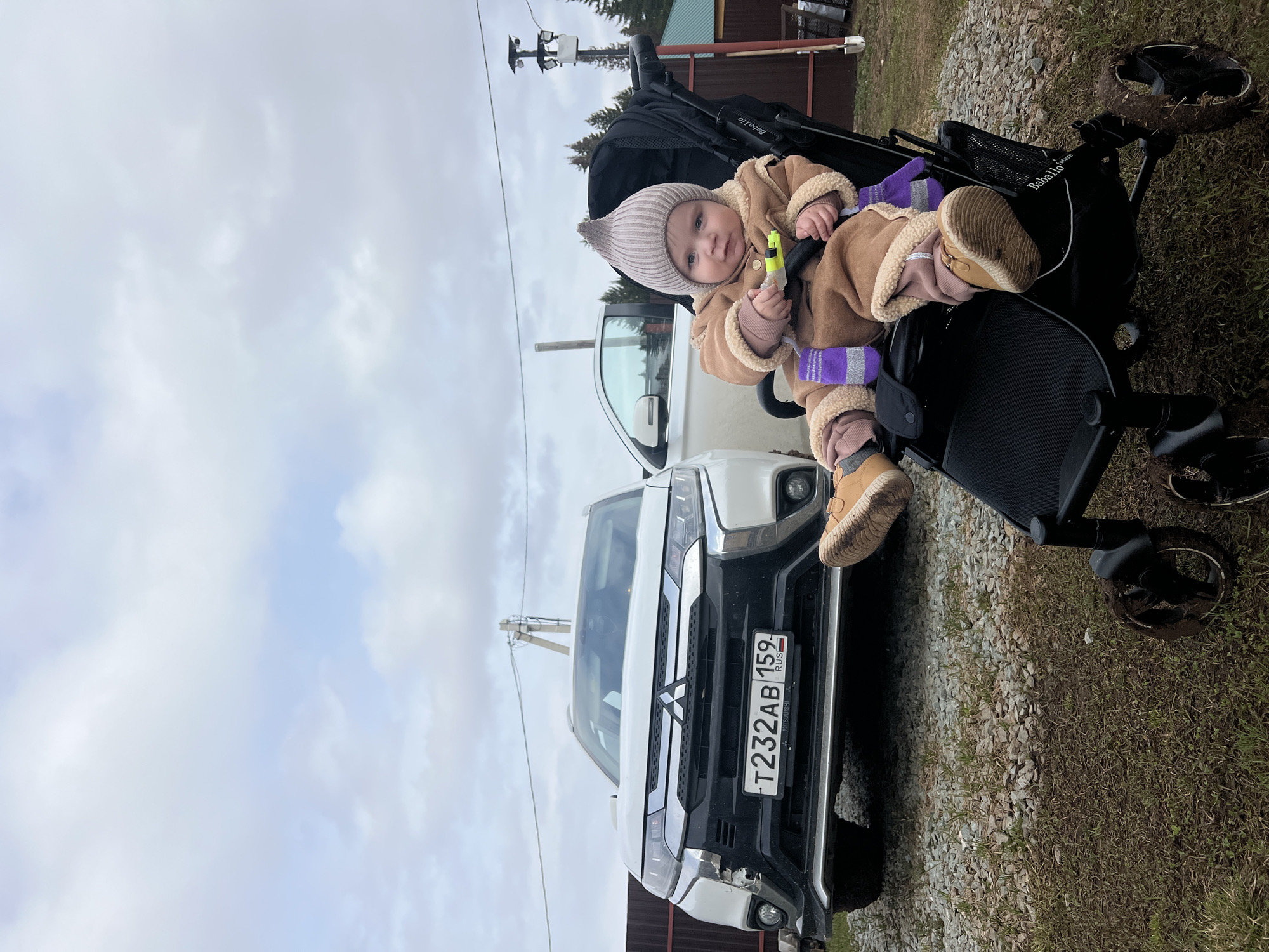 The image depicts a baby sitting in a black stroller. The baby wears a brown jacket with white fur trim, a light brown knitted hat, and brown shoes. The baby is holding a small yellow object. Behind the stroller is a white Mitsubishi Outlander with the license plate "T232AB 159 RUS", placing it in Russia. The car is parked on a gravel area next to a patch of grass. In the background, there's a wooden building with a dark brown roof, a power pole, and an overcast sky. The stroller appears to be a "Babaloo Future" model. This image was taken at a location near or in Protasy, Russia, as indicated by the license plate's regional code.