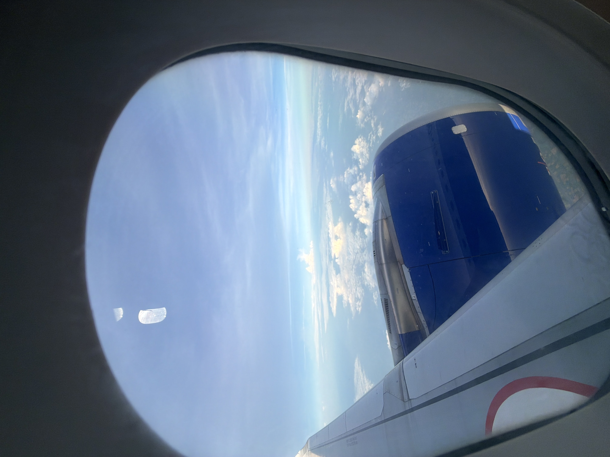 The image shows a view from an airplane window looking out at the sky and clouds. A portion of the airplane's wing and engine are visible in the foreground. The engine is blue and silver, and the wing is white with a red and white circular design near the bottom. The sky is a clear blue with scattered white clouds. The image does not contain any information or visual cues that would indicate it is located in Nadergul, India, or any other specific city.