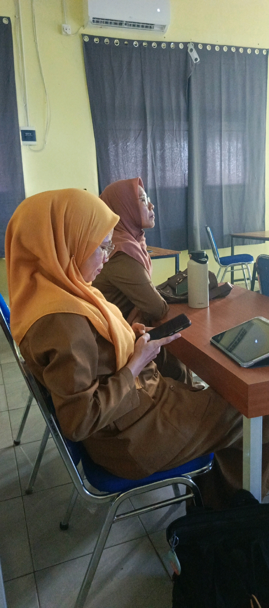 Two women wearing hijabs and brown uniforms are seated at a wooden table in what appears to be a classroom or meeting room. The woman in the foreground, wearing an orange hijab, is looking down at her phone. The woman behind her, wearing a pinkish-brown hijab, is looking forward. On the table are a water bottle, a bag, a tablet, and a mobile phone. The room has yellow walls, dark grey curtains, and a tiled floor. An air conditioning unit is mounted on the wall above the curtains. This scene is located in Desa Sarang Mandi, Indonesia.
