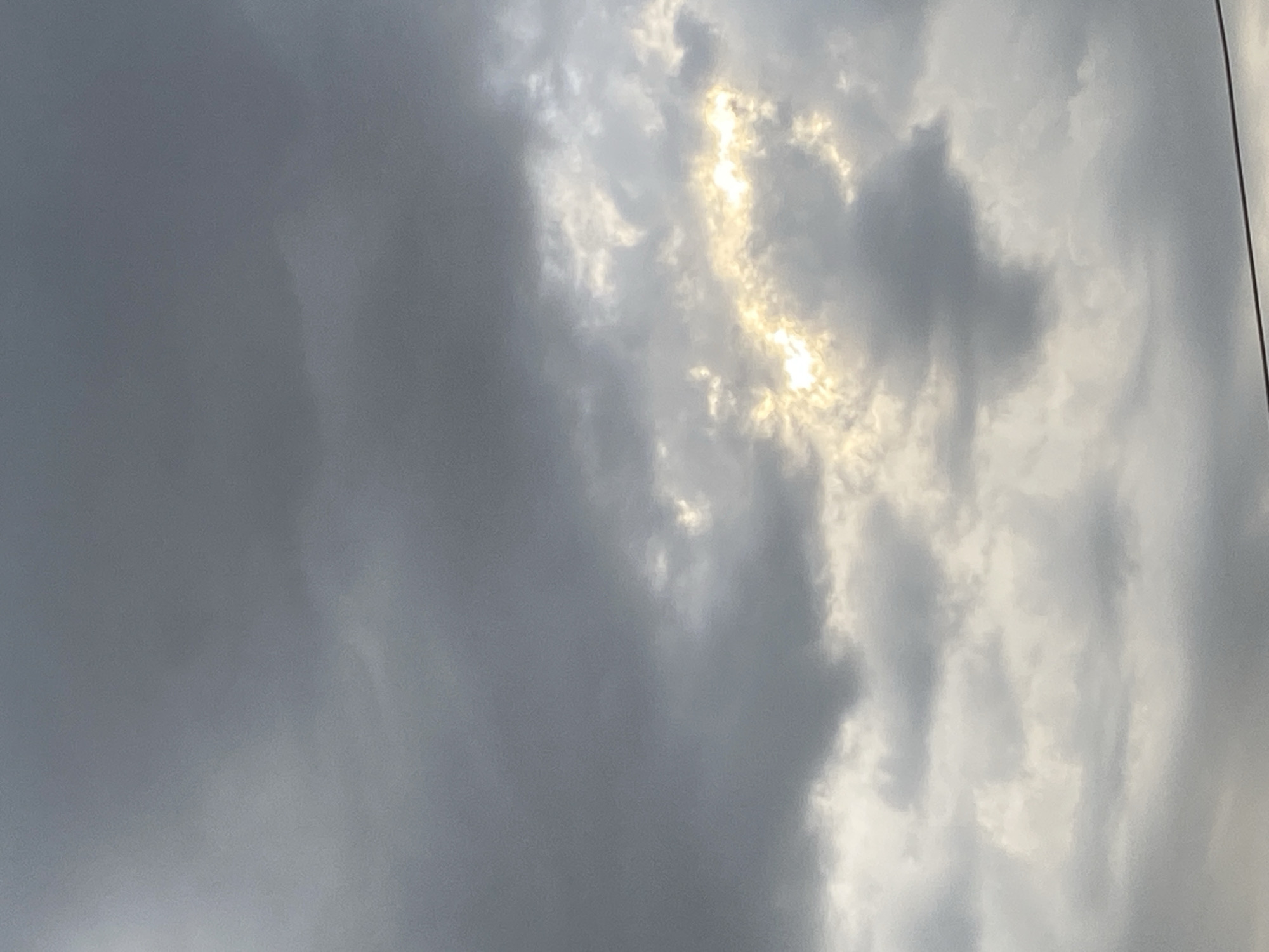 The image is a close-up, vertical shot of a cloudy sky above the city of Taygarayev, Kyrgyzstan. The clouds are predominantly dark gray, with some lighter, brighter areas where sunlight is breaking through. The sun's rays create a luminous, almost golden glow in a patch of the sky, highlighting the textured edges of the clouds. The overall mood is somewhat dramatic due to the contrast between the dark clouds and the shafts of light. There are no people, buildings, or distinct geographical features visible, only the atmospheric conditions. A thin, dark horizontal line, possibly a wire or cable, is visible across the bottom of the frame, suggesting a very low angle of the shot.