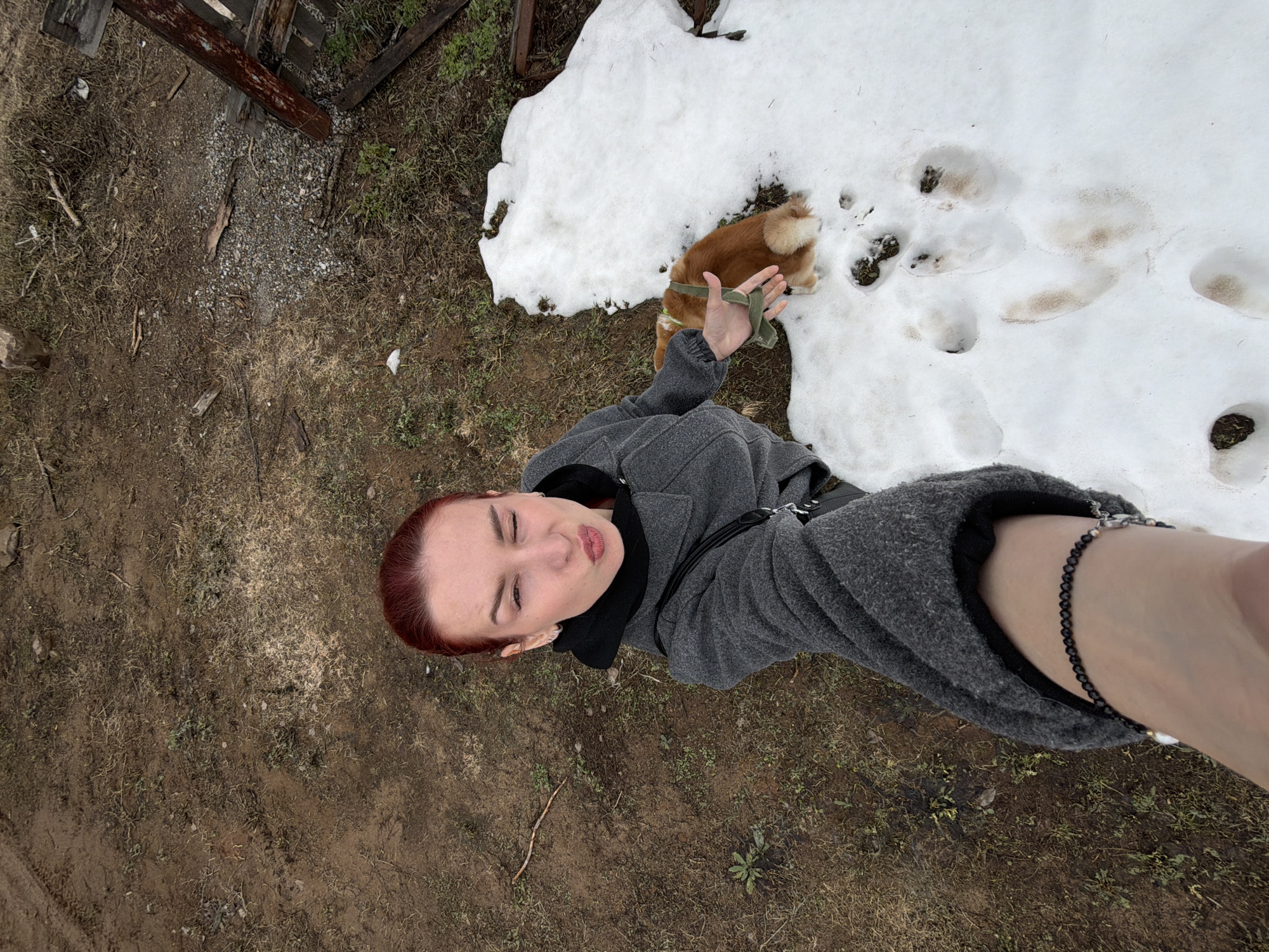 A woman is standing outdoors in what appears to be a yard or field near a building. The ground is mostly dirt and grass with some snow patches. A small dog is standing on the snow. The woman is looking at the camera with her lips pursed and has her arm extended towards the camera. The location is in the city of Chaikovsky, Russia.