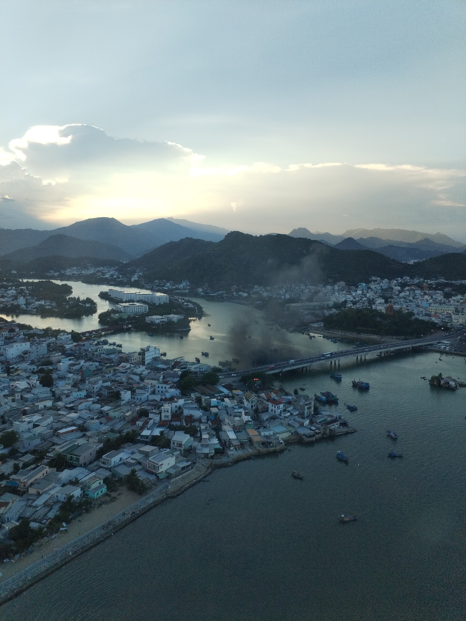 The image shows an aerial view of Bac Nha Trang Ward, Vietnam, with a landscape consisting of mountains, a river with a bridge, and residential areas. The river separates areas of densely packed buildings, with many having red tile roofs. Smoke rises from a point near the bridge in the middle ground. The bridge appears to have vehicular traffic on it. There are several boats scattered on the river and near the coast. The sky is overcast with light coming through breaks in the clouds above the mountains.