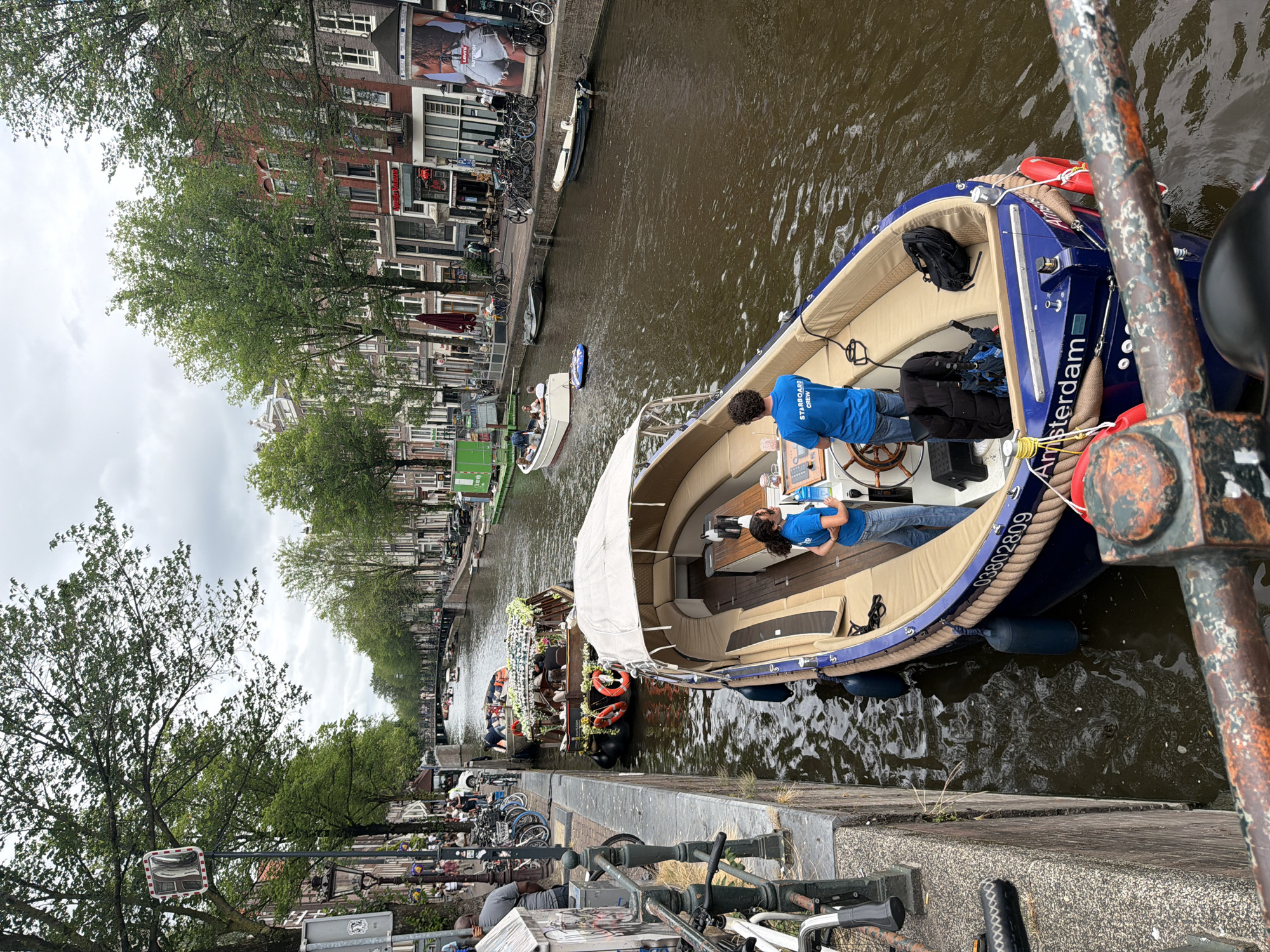 An overhead view captures a blue boat with "Amsterdam" written on its side, cruising through a canal in Amsterdam, Netherlands. Two people, appearing to be crew members, are visible on the boat. One, wearing a blue t-shirt and jeans, stands at the helm, while another, in a blue t-shirt, stands nearby. The boat is adorned with cream-colored cushions and a white canopy. Several other boats, including a larger, more ornate vessel decorated with flowers, are also present in the canal. The canal banks are lined with trees and buildings, characteristic of Amsterdam's architecture. The weather appears overcast.