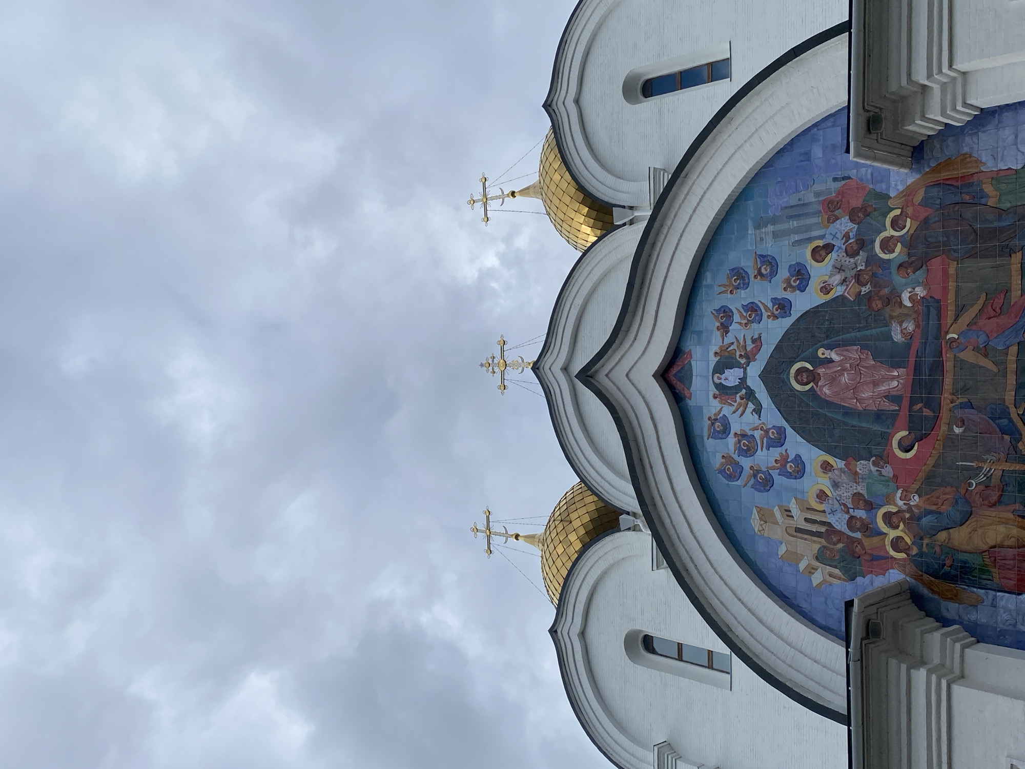 The image shows a close-up, upward view of the Church of the Icon of the Mother of God of All the Afflicted Joy, located in Шебунино, Russia. The architectural details include white walls, golden domes topped with Orthodox crosses, and a large semicircular mosaic depicting religious figures. The sky is overcast with grey clouds. No people are visible in the frame.