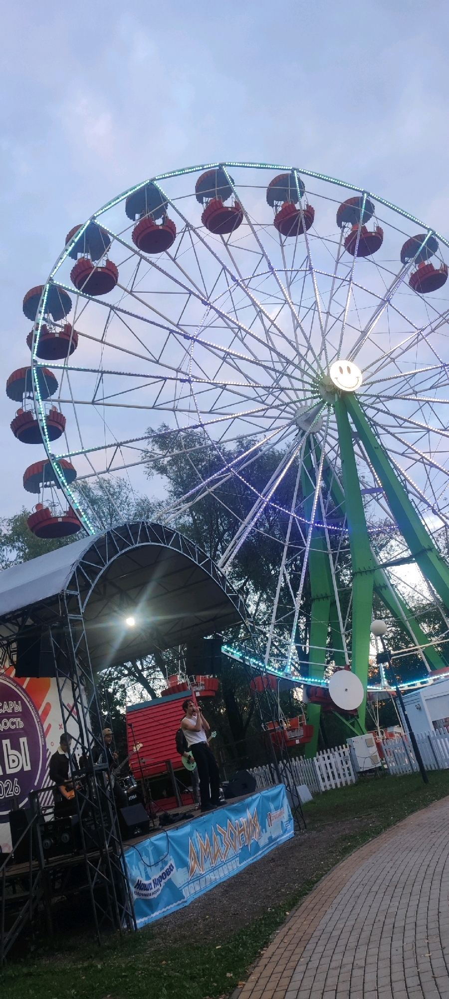 In Cheboksary, Russia, an outdoor concert is taking place in front of a Ferris wheel. A band performs on a stage set up beneath a covered structure. The lead singer, wearing a white tank top and black pants, is actively singing into a microphone. Other band members can be seen playing instruments. A large banner with blue and yellow graphics is displayed across the front of the stage, featuring text that appears to be in Russian and the word "AMAZONIA." Behind the stage, the illuminated Ferris wheel stands tall against a cloudy sky. The Ferris wheel has red passenger gondolas and is decorated with a string of white and blue lights, creating a festive atmosphere. The ground surrounding the stage is covered with gravel and grass, and a paved walkway is visible to the right. Trees are present in the background, suggesting a park-like setting.