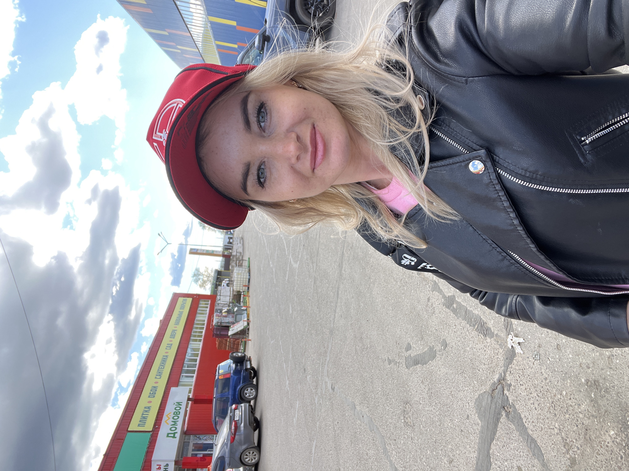 A woman with blonde hair and a red baseball cap is taking a selfie in front of a building. She's wearing a black leather jacket over a pink top. Behind her is a red building with a sign in Russian that reads "Домовой" (Domovoy), which translates to "House Spirit," a common name for home improvement or hardware stores in Russia. The sign also lists products such as "ПЛИТКА • ОБОИ • САНТЕХНИКА • САД • ДВЕРИ • НАПОЛЬНЫЕ ПОКРЫТИЯ," meaning "Tiles, Wallpaper, Plumbing, Garden, Doors, Flooring." There are cars parked in front of the building, and the sky is visible with some clouds. Based on the "Домовой" sign and the Cyrillic text, the location is likely Chaikovsky, Russia, as this is where the brand is known to be located.