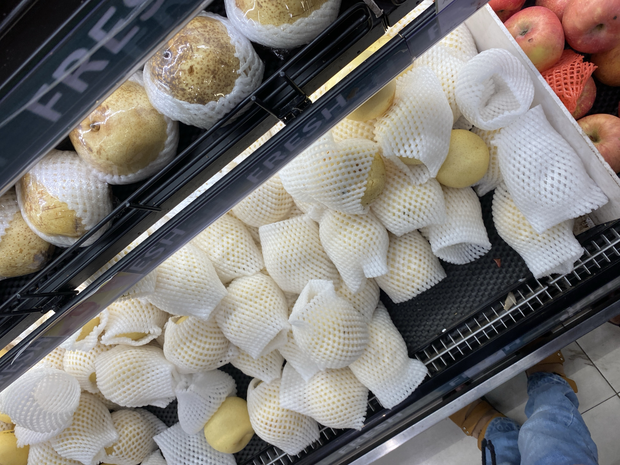 The image depicts a close-up view of a display of pears and apples at a market stall in Banda Aceh, Indonesia. The pears are individually wrapped in white protective netting. Some pears are a pale yellow color, while others are wrapped in plastic, revealing a yellow-brown skin with brown speckles. Apples in various shades of red and yellow are visible in a lower bin, some also partially covered by orange netting. The display is organized in metal tiered shelves. The background shows a glimpse of a person's legs, wearing blue jeans and brown sandals, suggesting a shopper is present. The overall environment appears to be an indoor market with a focus on fresh produce.