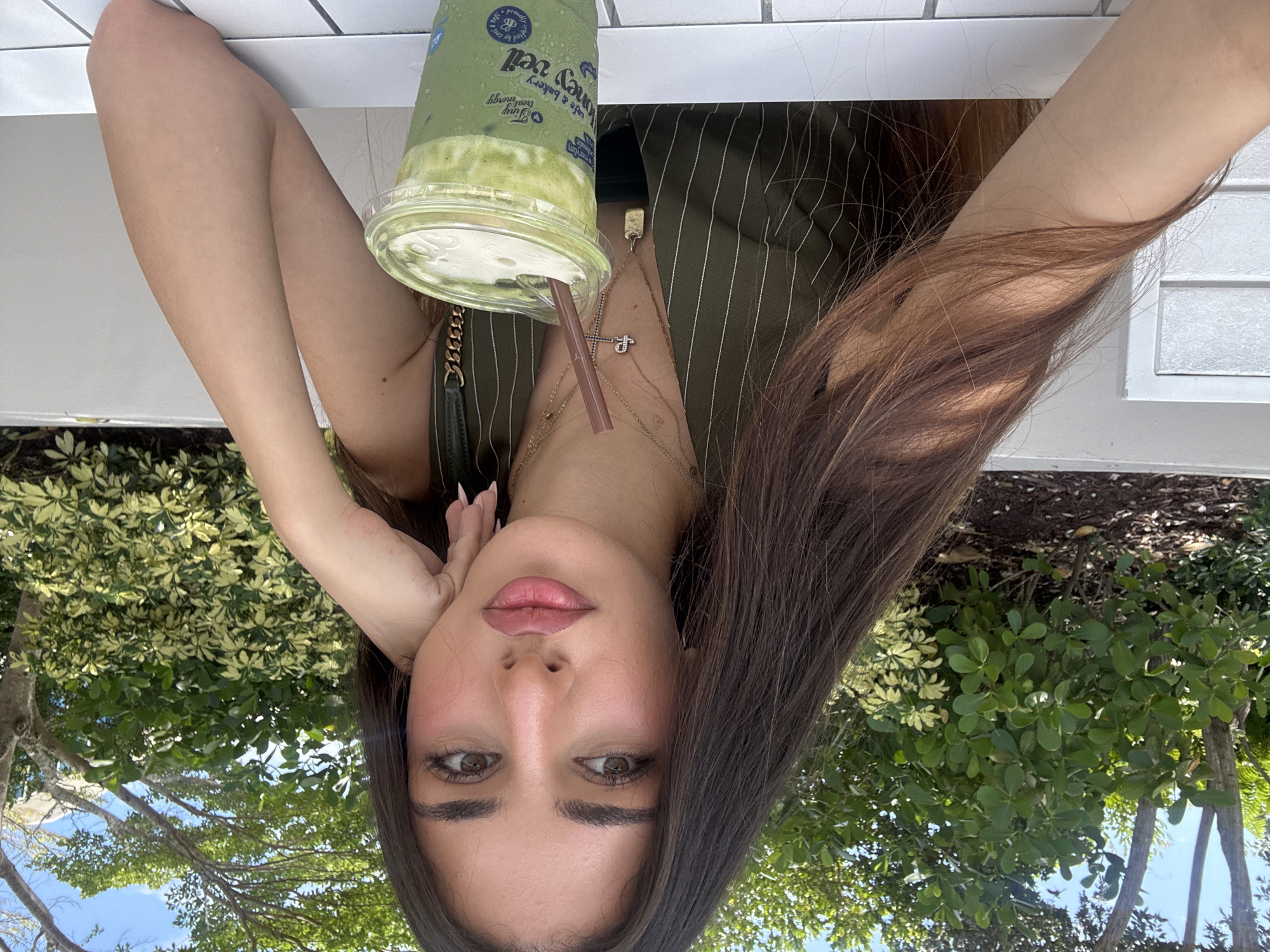 In Hallandale Beach, Florida, USA, a young woman with long, dark brown hair is seated outdoors at a white table, captured in a medium shot. She is leaning her head on her left hand, with her arm resting on the table. The woman is wearing a dark green pinstripe vest with a low-cut neckline, adorned with several necklaces, including a pendant with a cross. A clear plastic cup filled with a green beverage, likely a matcha latte, with a brown straw, sits on the table to her right. Behind her, lush green foliage and trees are visible under a clear blue sky, suggesting a bright, sunny day. The background also shows hints of outdoor seating and a textured white wall.