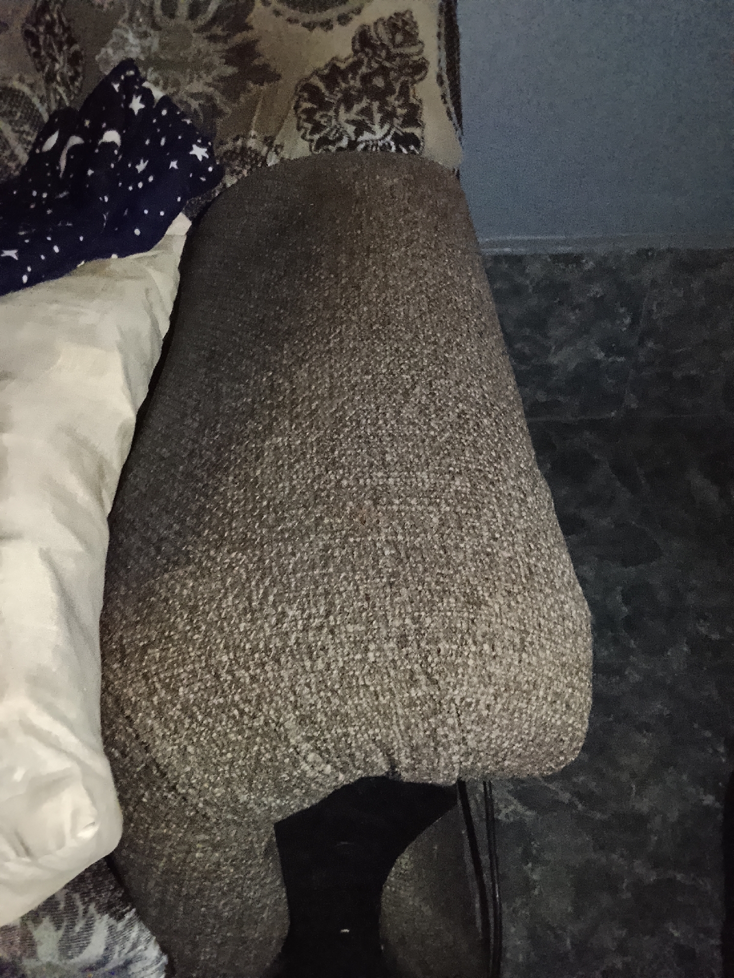 The image shows a close-up view of a recliner chair, upholstered in a textured brown fabric. A white pillow and a dark blue blanket with white stars and moons are draped over the arm of the chair. The background includes a patterned upholstery on what appears to be another piece of furniture, and a dark, textured surface that could be a wall or floor. The lighting is dim, suggesting an indoor setting, possibly at night or in a dimly lit room. There are no people visible in the image, and no text is present. The scene is static and focuses on the textures and colors of the domestic objects.