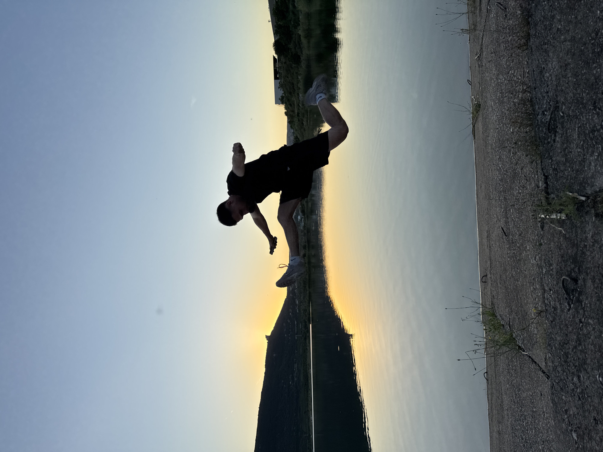 A silhouette of a young man is captured mid-air, performing a split leap over a body of water at sunset. He is wearing a dark t-shirt and shorts, with gray athletic shoes. His body is horizontal in the air, with his left leg extended forward and his right leg extended backward. His arms are outstretched, balancing his pose.
The background features a serene body of water reflecting the warm hues of the setting sun, which is partially obscured by a dark, silhouetted hill on the left side of the frame. The sky above is a gradient of soft blues and yellows, indicating either early morning or late evening. On the right side of the water, there is a verdant shoreline with some buildings visible in the distance. The foreground consists of a rough, concrete surface with patches of dry grass and scattered debris, suggesting a man-made embankment.
This scene is likely near the Shumen Plateau, in the vicinity of the Shumen Reservoir, which is a popular recreational area known for its natural beauty and walking paths. The reservoir is located to the west of the city of Shumen, Bulgaria.
