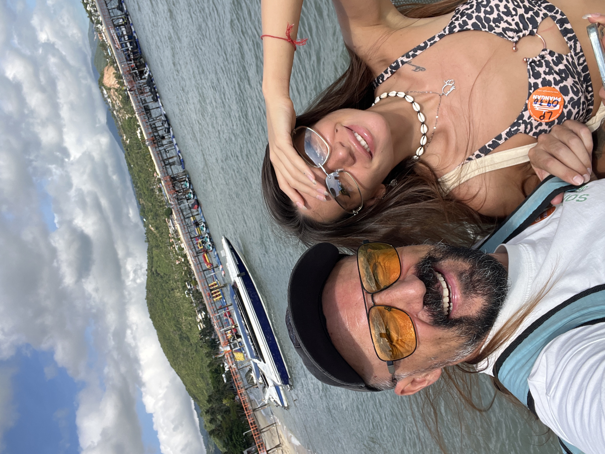 In Baan Bang Rak, Thailand, on a bright, cloudy day, a man and a woman take a selfie by the water. The man, wearing sunglasses with orange lenses and a black baseball cap, smiles broadly, his beard and hair blowing in the wind. He has a backpack on. The woman, with long brown hair, wears sunglasses and a leopard-print bikini top adorned with a seashell necklace and a red string bracelet. Her hand is raised to her forehead, and she smiles. In the background, a sleek white and blue motorboat is docked near a pier with numerous jet skis. The pier stretches out over the water, with buildings and lush green hills visible on the shore beyond. The sea is a calm, greyish-blue.