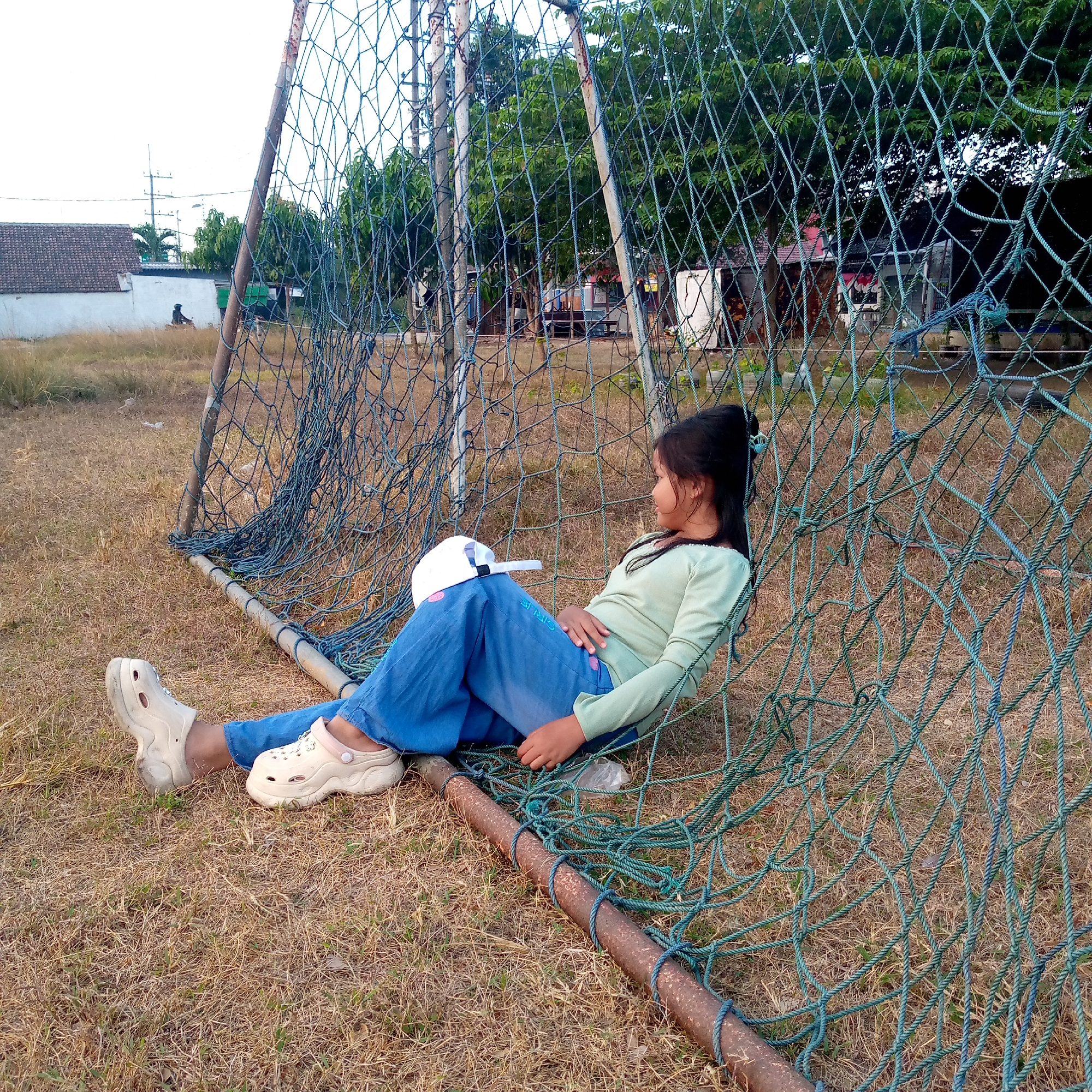 A young girl is sitting on the ground next to a rusty metal soccer goal, with her legs crossed and resting on a blue net. She is wearing blue jeans and a light green long-sleeved shirt, and white clogs. A white baseball cap is placed on her lap. The soccer goal is old and weathered, with a blue net that appears to be somewhat torn and frayed in places. The ground around the goal is covered in dry, yellowish grass. In the background, there is a white building with a grey tiled roof, some trees, and power lines. The sky is overcast. The image appears to have been taken in Pacing, Indonesia.