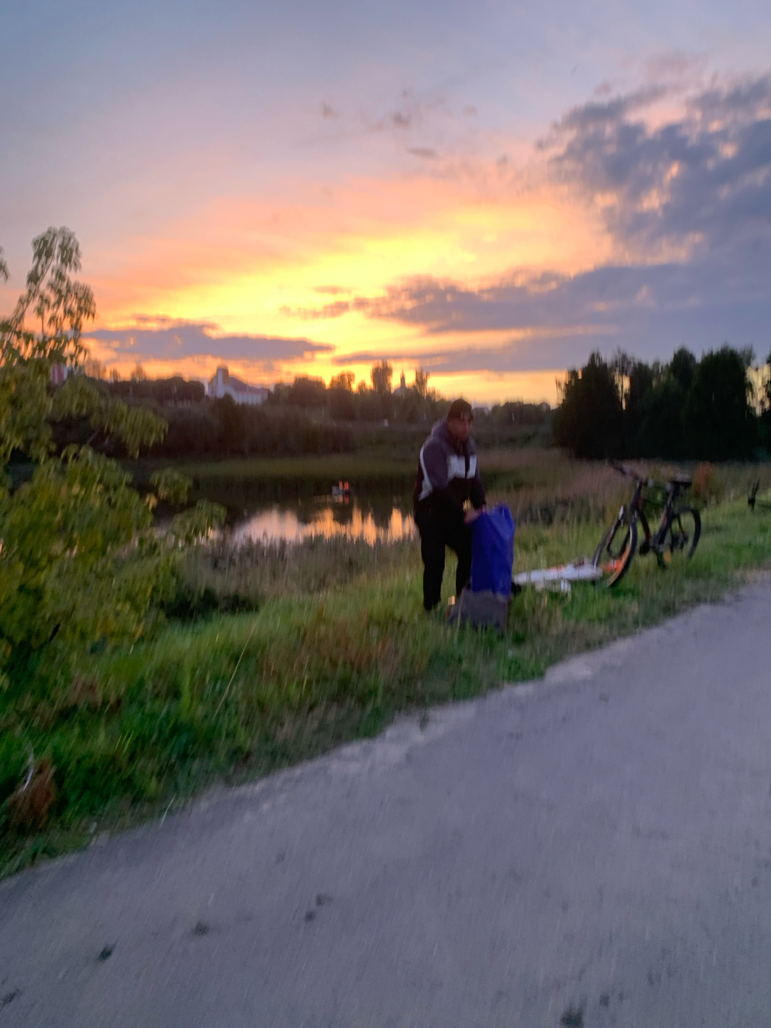 A person is packing a blue backpack next to a bicycle on the grassy bank of a body of water at sunset. The sky is filled with vibrant orange and yellow hues, with scattered clouds. In the distance, trees and buildings are visible, and the water reflects the sky. The overall scene suggests a moment of rest or preparation for departure in a natural setting near the city of Kapyĺ, Belarus.