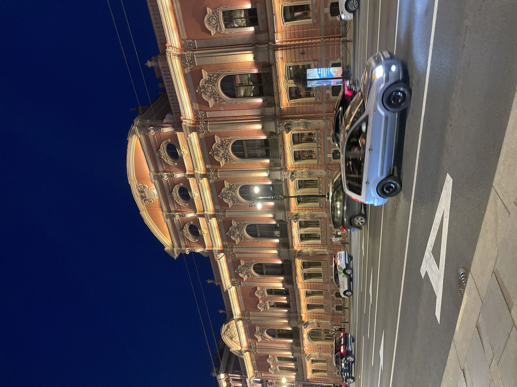 The image presents a nighttime street scene featuring the illuminated Neo-Baroque facade of the Beloselsky-Belozersky Palace on Nevsky Prospekt in Saint Petersburg, Russia. The multi-story palace, predominantly salmon-pink, is extensively illuminated by warm-toned floodlights that highlight its intricate architectural details, including numerous arched and oval windows with ornate surrounds, decorative pilasters, and a prominent curvilinear pediment at the roofline. Large sculpted male figures, identified as atlantes, are visible supporting the upper stories along the lower section of the building's facade.
In the foreground, a wide, multi-lane asphalt road displays white painted directional arrows. A light blue Ford Fusion-type hatchback, showing significant motion blur on its body and wheels, moves from right to left with its headlights on. Other vehicles, including a dark sedan, a white SUV, and a dark SUV, are also in motion further down the road. A paved sidewalk, visible in the lower left, features a metal manhole cover. An advertising pillar with an illuminated poster, displaying text in Russian, stands on the sidewalk directly in front of the palace. A classical black lamppost with a glowing spherical fixture is also present. The sky above the building is a deep, dark blue, indicating late evening or night.