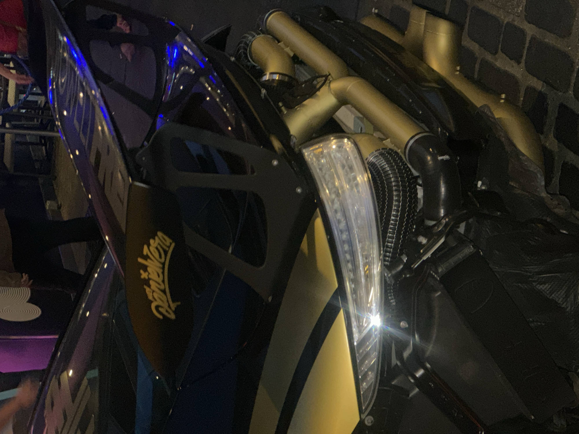 The image shows a black and gold vehicle, partially visible, with the word "Pantera" written on the side. The vehicle is parked near a brick wall. Various gold pipes and black hoses are visible. The environment appears dimly lit, and people are partially visible in the background.