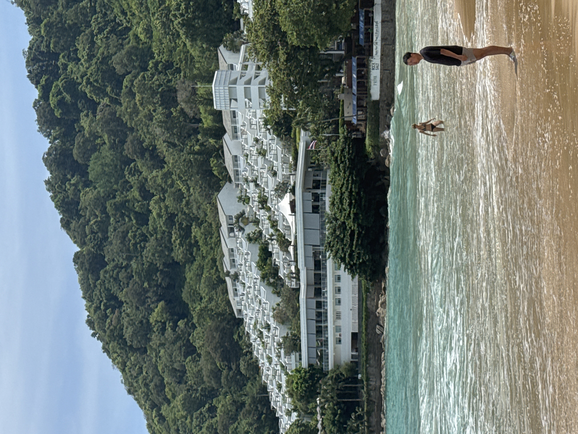 The image depicts a tropical beach scene in Rawai, Thailand, likely at a resort. A man in a black t-shirt and grey shorts stands on the sandy shore, facing the ocean. Further out in the shallow, turquoise water, a woman in a black bikini is wading. The ocean exhibits moderate waves with white foam. In the background, a multi-tiered white resort complex is built into a lush, green hillside covered with dense tropical foliage. The resort features numerous balconies and white structures ascending the slope, with a larger building at the base. The sky is clear and bright blue.
