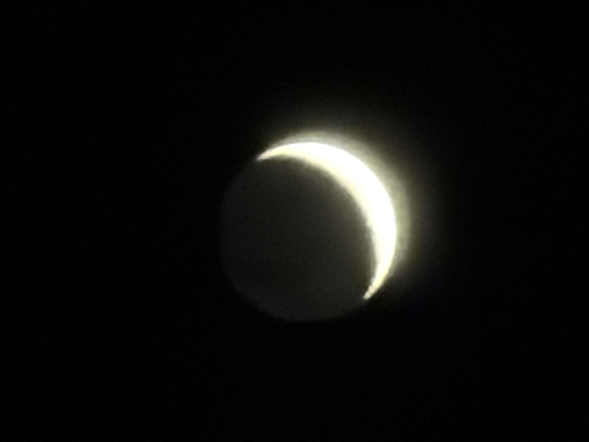 A crescent celestial body, likely the Moon or Venus, is positioned centrally against a black background. Only a small sliver of the orb is illuminated, exhibiting a bright, white-yellow hue. The remaining portion of the orb appears dark, though with a subtle, ghostly luminescence, suggesting faint earthshine or atmospheric scattering. The image lacks the presence of any landmarks or contextual cues to definitively establish a location. Therefore, it's impossible to confirm that it shows London, United Kingdom.