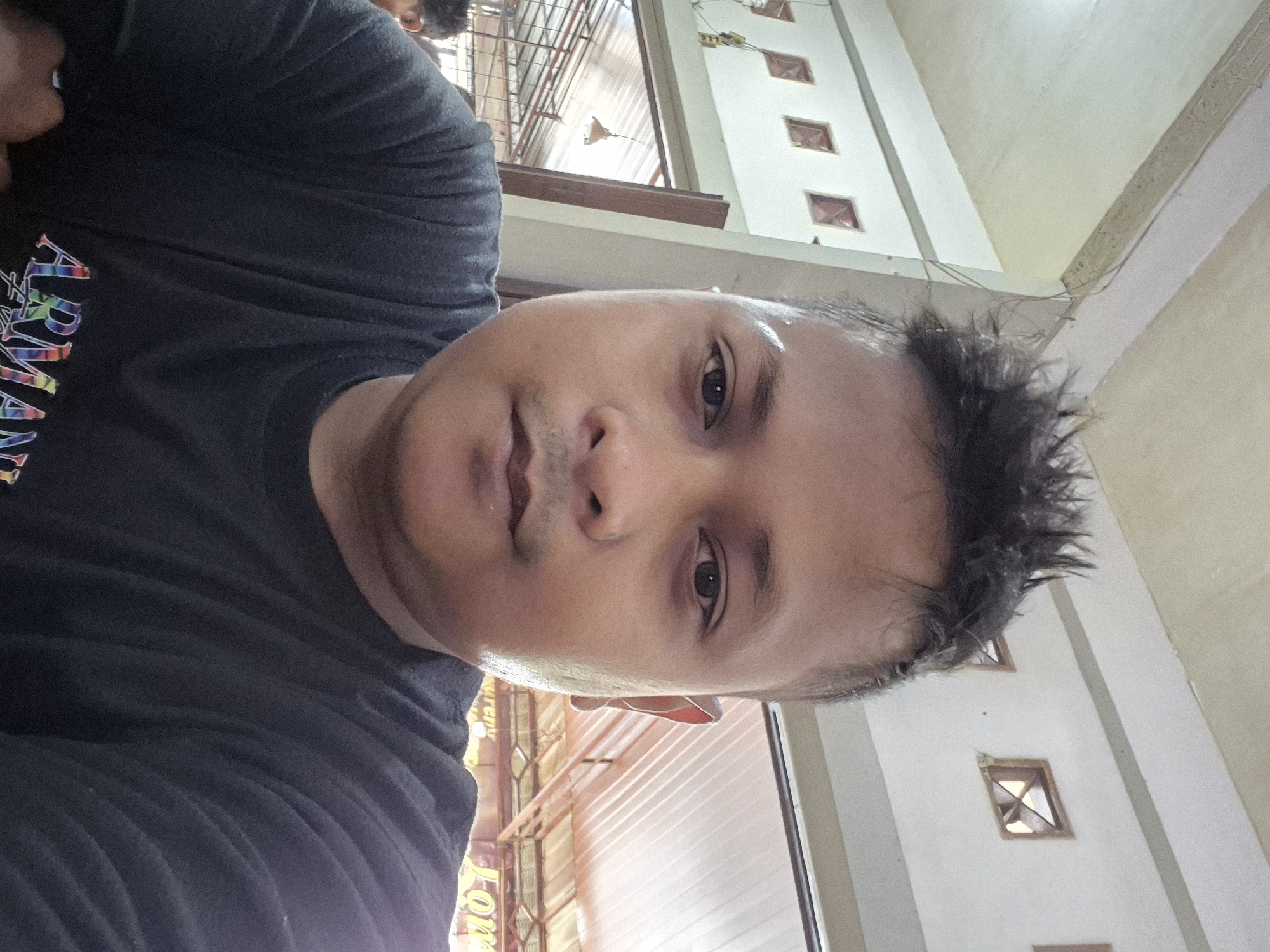 A selfie-style image captures a man's face and upper torso, taken in what appears to be an indoor or semi-indoor public space in Lhokseumawe, Indonesia. The man, with dark, short, spiky hair and light stubble, looks directly at the camera with a neutral expression. His face, particularly his forehead, appears to be glistening with sweat, suggesting a warm or humid environment.
He is wearing a black t-shirt, on which the brand name "ARMANI" is clearly visible on the left side of his chest, rendered in colorful, stylized letters. The background shows light-colored walls, possibly white or cream, featuring a series of small, square, recessed openings that could be windows or decorative elements. Above, a ceiling structure with exposed beams or trusses is visible, along with a hanging light fixture. A partial view of another person's head and shoulder can be seen in the distant background. The natural light suggests the photo was taken during the daytime. Partial text, "LOJ...", is faintly discernible on an object in the lower-left background, but is too incomplete for identification.
