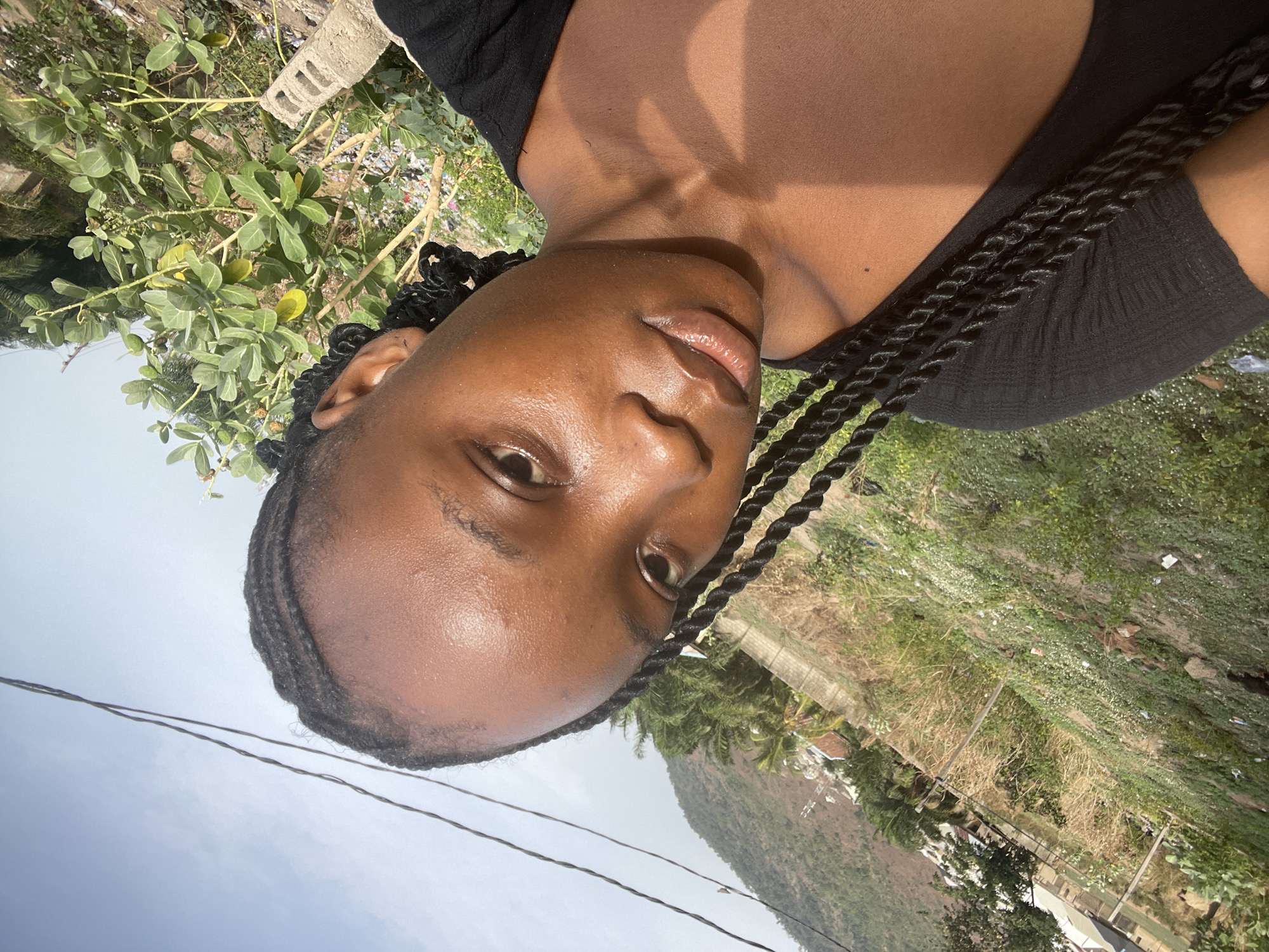 The image is a close-up, upward-facing portrait of a young woman with dark skin. Her hair is styled in numerous small, dark braids (cornrows) that frame her face and extend over her left shoulder. She has dark eyes and looks directly into the camera with a neutral expression. She is wearing a dark, likely black, scoop-neck top. Direct sunlight illuminates her face and upper body, casting distinct shadows on her neck and chest, indicating a bright and clear day.
The background depicts an outdoor scene. Immediately behind her, there is dense green foliage mixed with some dry, lighter-colored vegetation, and a concrete block is partially visible. Further in the mid-ground, several distant buildings and structures are discernible amidst more trees and greenery. A prominent, tree-covered mountain or large hill dominates the distant horizon under a pale blue, clear sky. Two dark power lines stretch across the upper portion of the sky. The setting suggests a semi-urban or rural environment, consistent with the provided location context of Lokoja, Nigeria. No visible text is present in the image. The time of day is daytime, characterized by strong sunlight.