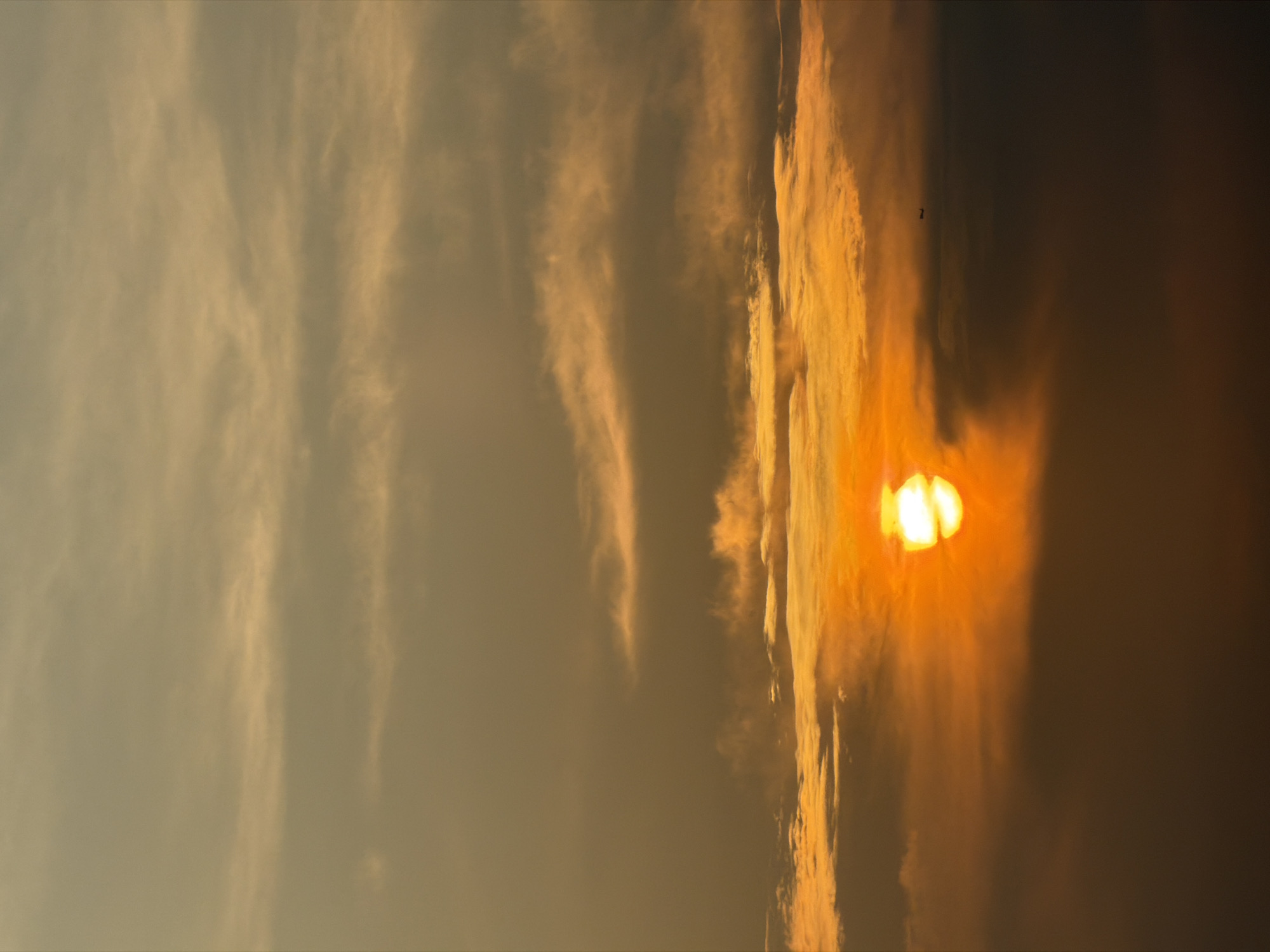The image captures a dramatic sunset over the city of Jeju, South Korea. The sun, a bright orb of orange and yellow, is partially obscured by dark, brooding clouds that create a stark contrast with the fiery sky. Wisps of lighter clouds drift higher, tinged with the golden hues of the setting sun. In the distance, a small silhouette of what appears to be an airplane can be seen against the hazy horizon. The lower portion of the image is dominated by darker clouds, giving a sense of depth and drama to the scene.
