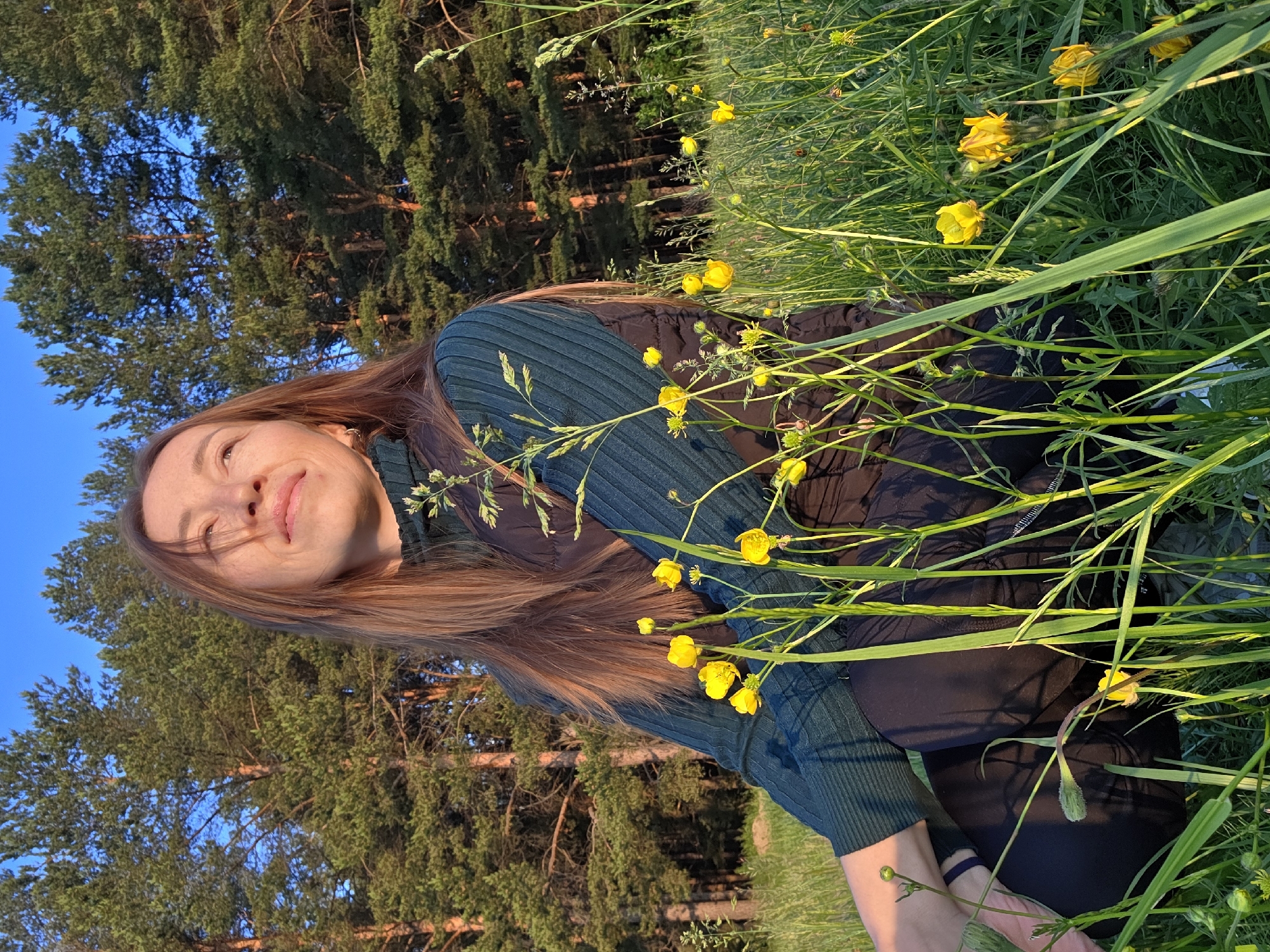 A young woman with long, reddish-brown hair is captured in a horizontal outdoor shot, smiling gently and looking upwards. She is dressed in a dark teal or green ribbed long-sleeved top and dark pants. Her face shows faint freckles, illuminated by warm, golden light.
The woman is positioned within a vibrant natural landscape, surrounded by tall green grass and numerous bright yellow buttercup flowers. In the background, a dense line of pine trees stands tall against a clear blue sky. The lighting suggests late afternoon or early evening, often referred to as the "golden hour," casting soft, warm tones across the scene and creating distinct shadows.
The woman appears relaxed and content, enjoying the serene environment. The overall mood of the image is peaceful and joyful, set in a sunny, clear weather condition. There is no visible text within the media. The location context for this scene is Chaikovsky, Russia.