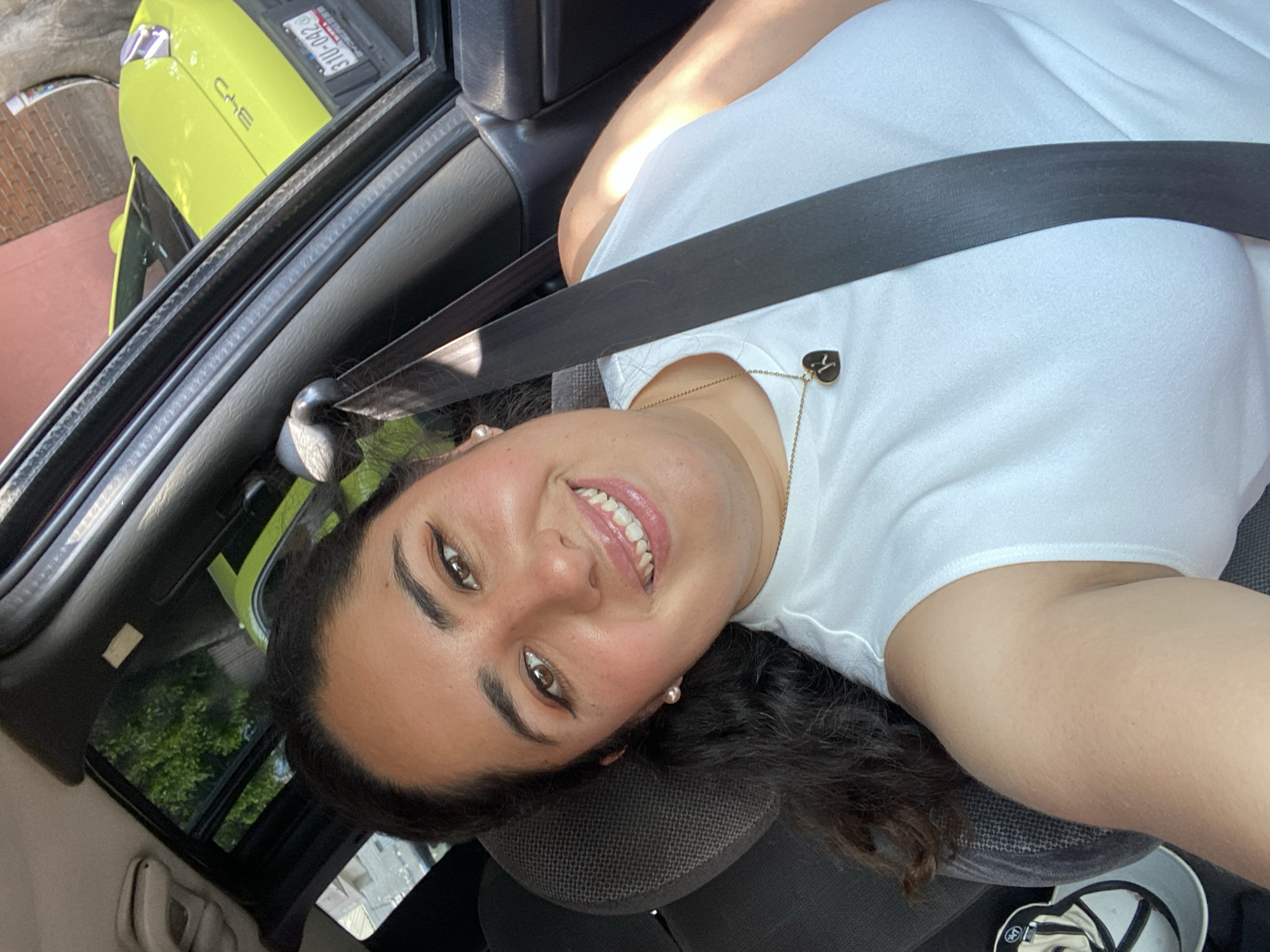 A woman is seated in a car, wearing a white shirt and a seatbelt. She is smiling and facing the camera, which is positioned at an angle above her. She wears a gold necklace with a heart-shaped pendant. A second vehicle, painted light green, is partially visible through the car window. The license plate number visible on the green vehicle is "311-D9Z". Foliage is visible through another window in the vehicle.
Based on the license plate information of the light green car, and assuming the car is registered in Mexico, the location of this photo is likely within Mexico, though not necessarily in Puebla. There is insufficient evidence to determine whether the photograph was taken in Puebla based on the contents of this image.