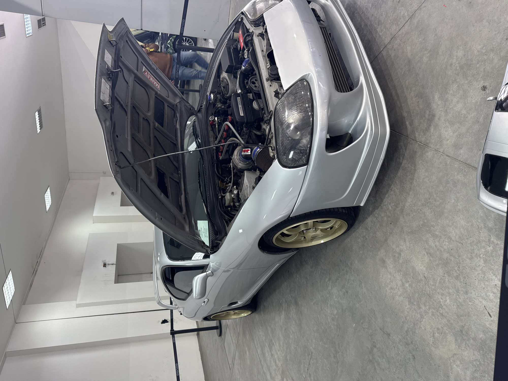 The image shows a silver car with its hood open, revealing the engine. The car is likely a Toyota Supra based on the engine layout. The surrounding environment is a well-lit indoor space with white walls and a gray floor, resembling a showroom or garage. There are other vehicles visible in the background, suggesting an automotive event or dealership. A person is partially visible near the open hood, possibly inspecting the engine. The location is not discernible as Plovdiv, Bulgaria from the visual information provided.