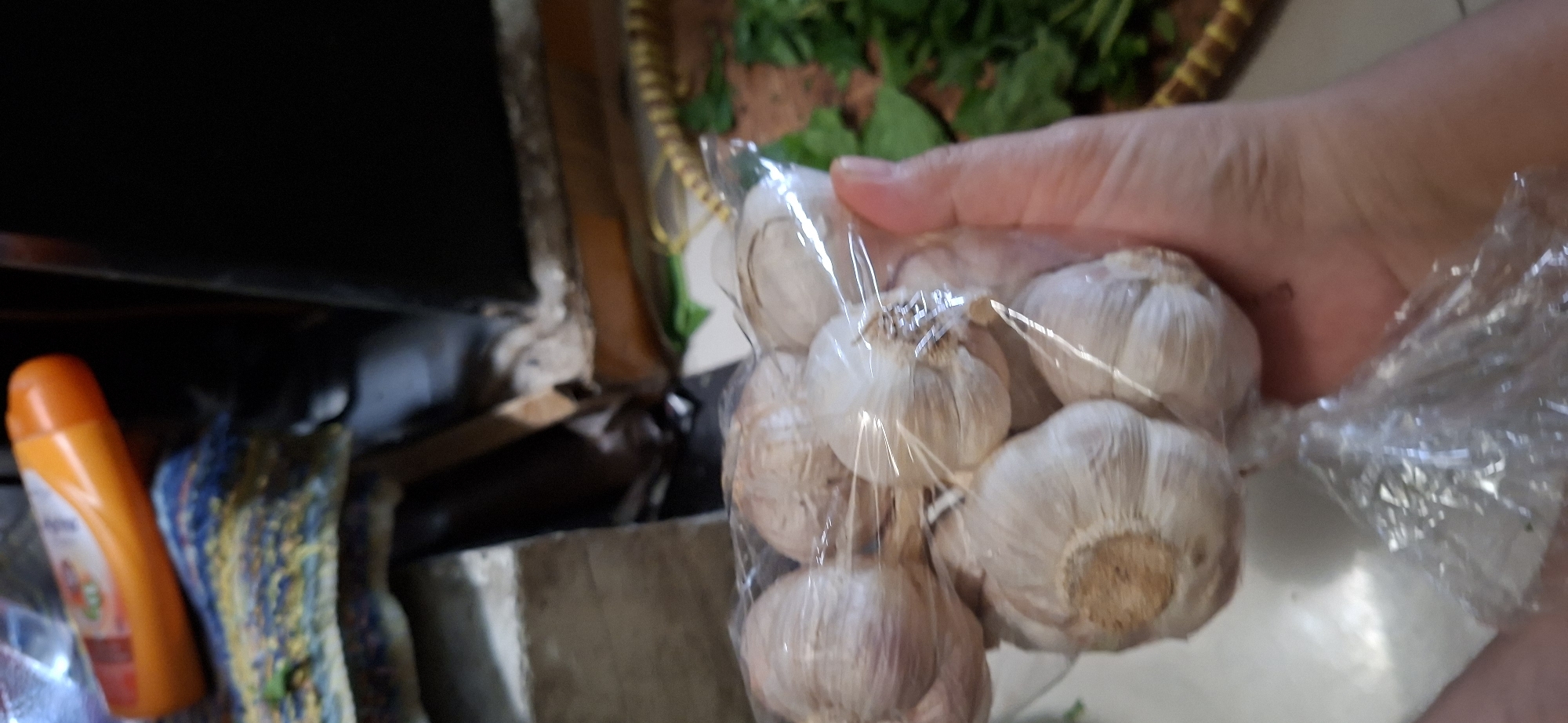 A person's hand is holding a clear plastic bag filled with several bulbs of garlic. The garlic bulbs are off-white in color with a slightly woody texture and are tied together with string. In the background, out of focus, is an orange bottle lying on a patterned blue and yellow fabric. To the right of the garlic, a bunch of green leafy vegetables can be seen. The scene appears to be indoors, likely in a residential setting in Surabaya, Indonesia, given the context of a food preparation or shopping scenario.
