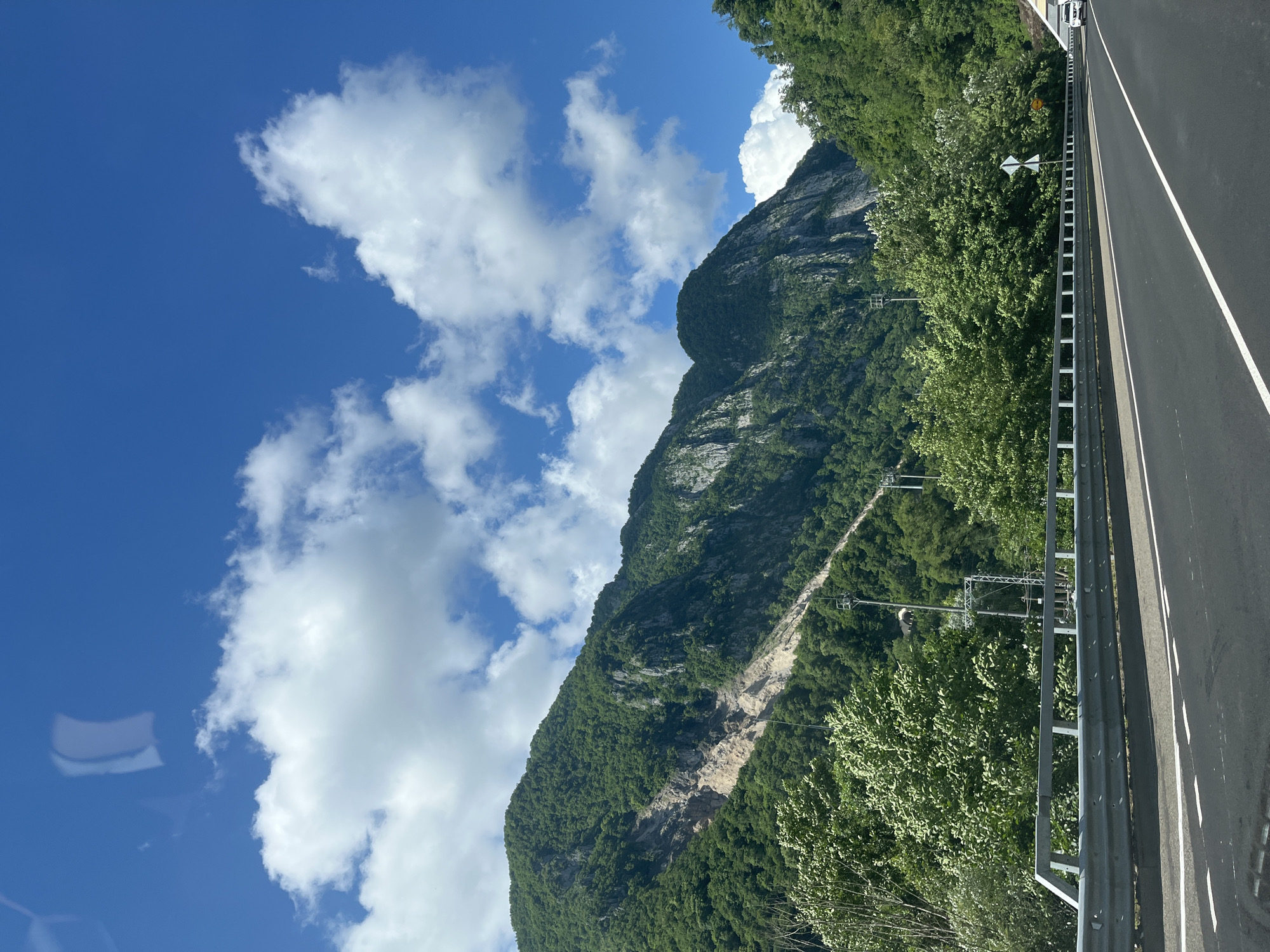 The image shows a landscape view from a road, likely a highway, in Monastyr, Russia. A mountain range dominates the background, with lush green trees covering most of its slopes. A significant scar of lighter-colored earth, indicative of a landslide or erosion, is visible on the side of one of the mountains. The sky is a vibrant blue, dotted with fluffy white clouds. In the foreground, a paved road stretches into the distance, with a metal guardrail separating it from the surrounding vegetation. There are no people clearly visible in the image. The environment suggests a natural, mountainous region.