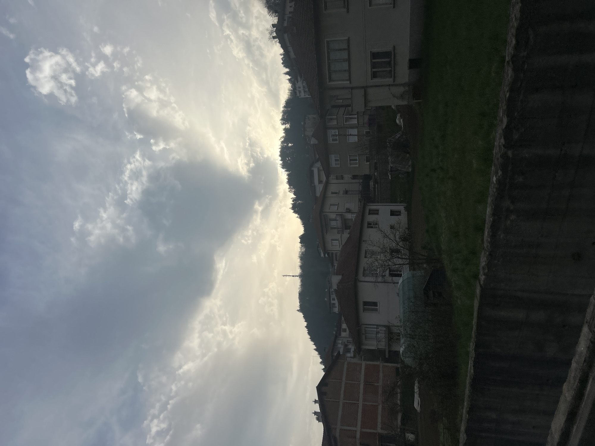 The image shows a view in Zlatograd, Bulgaria, looking towards a hillside with houses built along its slope. The sky is filled with clouds, and the sun appears to be behind them, creating a soft, diffused light. A tower can be seen amongst the trees in the distance. A stone wall is on the right side of the picture, and a patch of grass is visible above it.