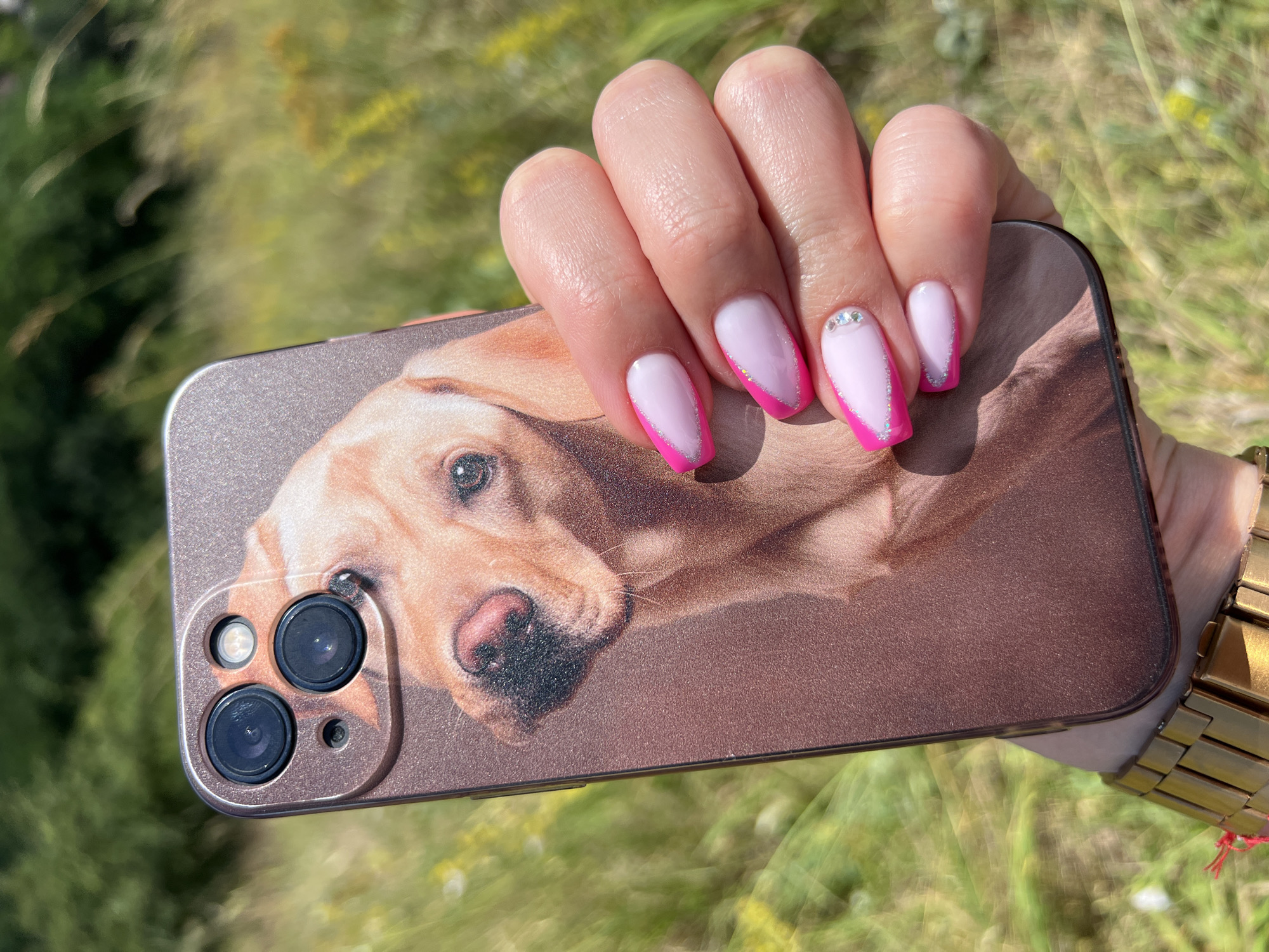 A close-up shot shows a person's right hand holding a smartphone with a case featuring a printed image of a golden retriever's face. The phone is positioned in front of a blurry background of green grass and foliage, suggesting an outdoor setting. The person's nails are manicured with a French tip design, featuring pink and white polish with a subtle glitter accent. A gold-colored wristwatch is visible on the person's wrist. The image appears to have been taken in Karniowice, Poland, based on contextual information.