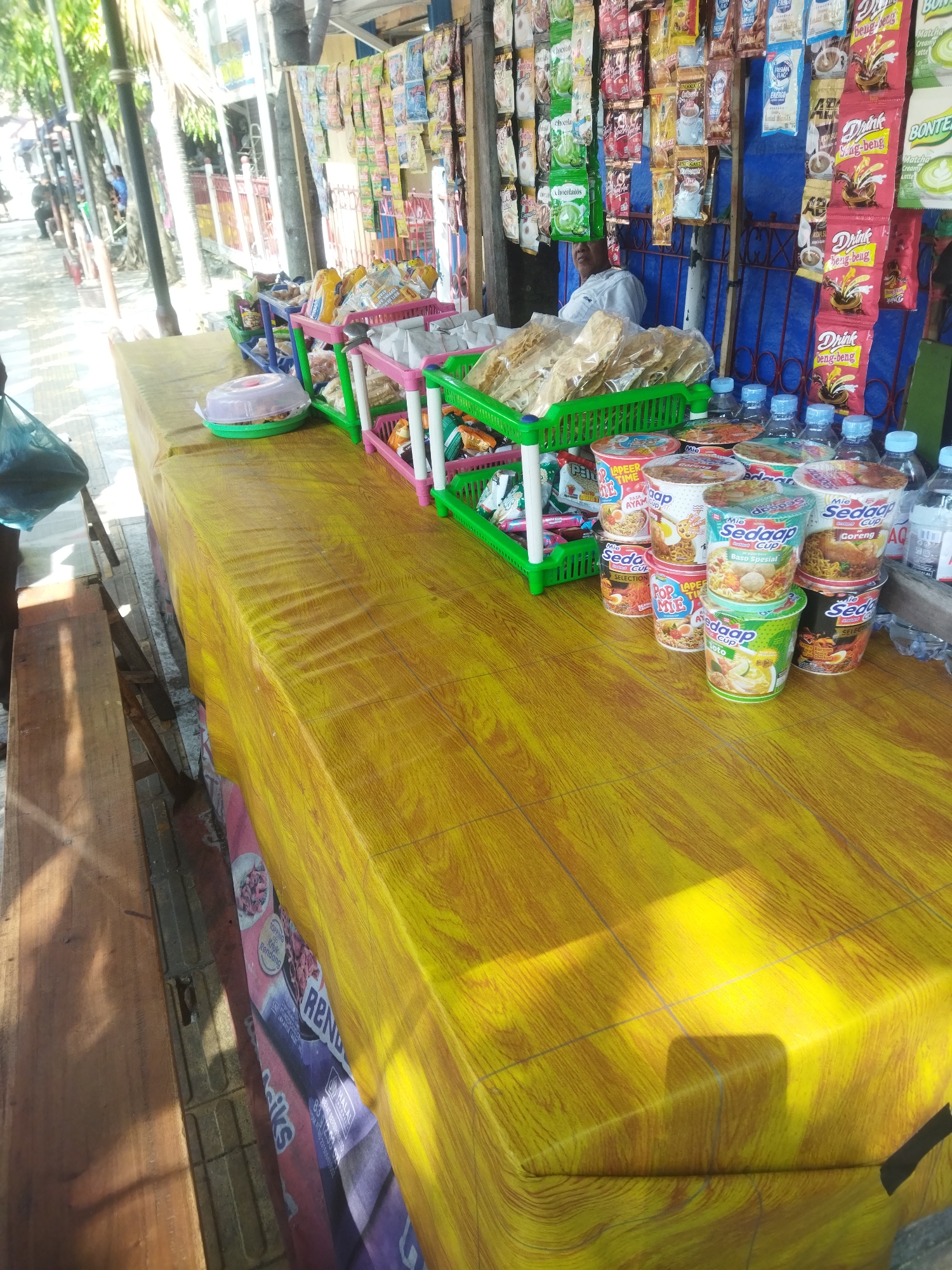 A street vendor stall in the city of Wonogiri, Indonesia, is set up on a yellow-patterned, wooden-look tablecloth. The stall displays an assortment of food and beverage items, including instant noodles in cups, packaged snacks, and bottled drinks. Several green, pink, and blue plastic baskets are filled with various snacks. In the background, a blue tarpaulin serves as a backdrop, with rows of packaged instant drinks and coffee hanging from a display rack. A person in a white shirt is partially visible behind the display. The stall is situated on a sidewalk, with a wooden bench visible to the left and other indistinct structures and trees in the background. The lighting suggests it is daytime, with some shadows cast on the tablecloth.