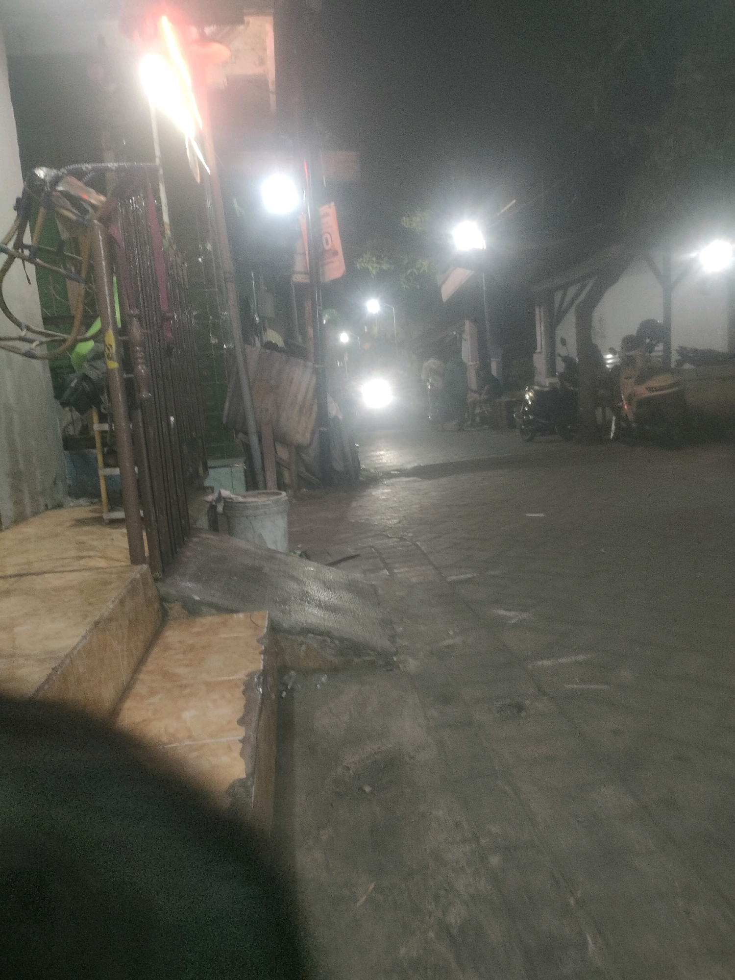 The image captures a dimly lit street scene at night, likely in Surabaya, Indonesia. In the foreground, a tiled stairway with a rough concrete edge is visible on the left. A dark, out-of-focus object occupies the lower left portion of the frame.
To the right of the stairway, a metal fence or gate with vertical bars runs along the sidewalk. Behind the fence, various objects are present, including a ladder, some bags, and a large metal bucket. Beyond this, the street extends into the background.
Several streetlights are illuminated, casting a bright glow that makes some areas appear overexposed. A motorcycle with its headlight on is moving down the street from the background towards the viewer, its light creating a strong beam. Other motorcycles and possibly people are dimly visible further down the street, some near small structures that appear to be shops or stalls. The road surface is paved with what looks like cobblestones or interlocking bricks. Trees with foliage are silhouetted against the night sky in the background. The overall impression is a bustling street at night, possibly a commercial area or a residential neighborhood with local businesses.