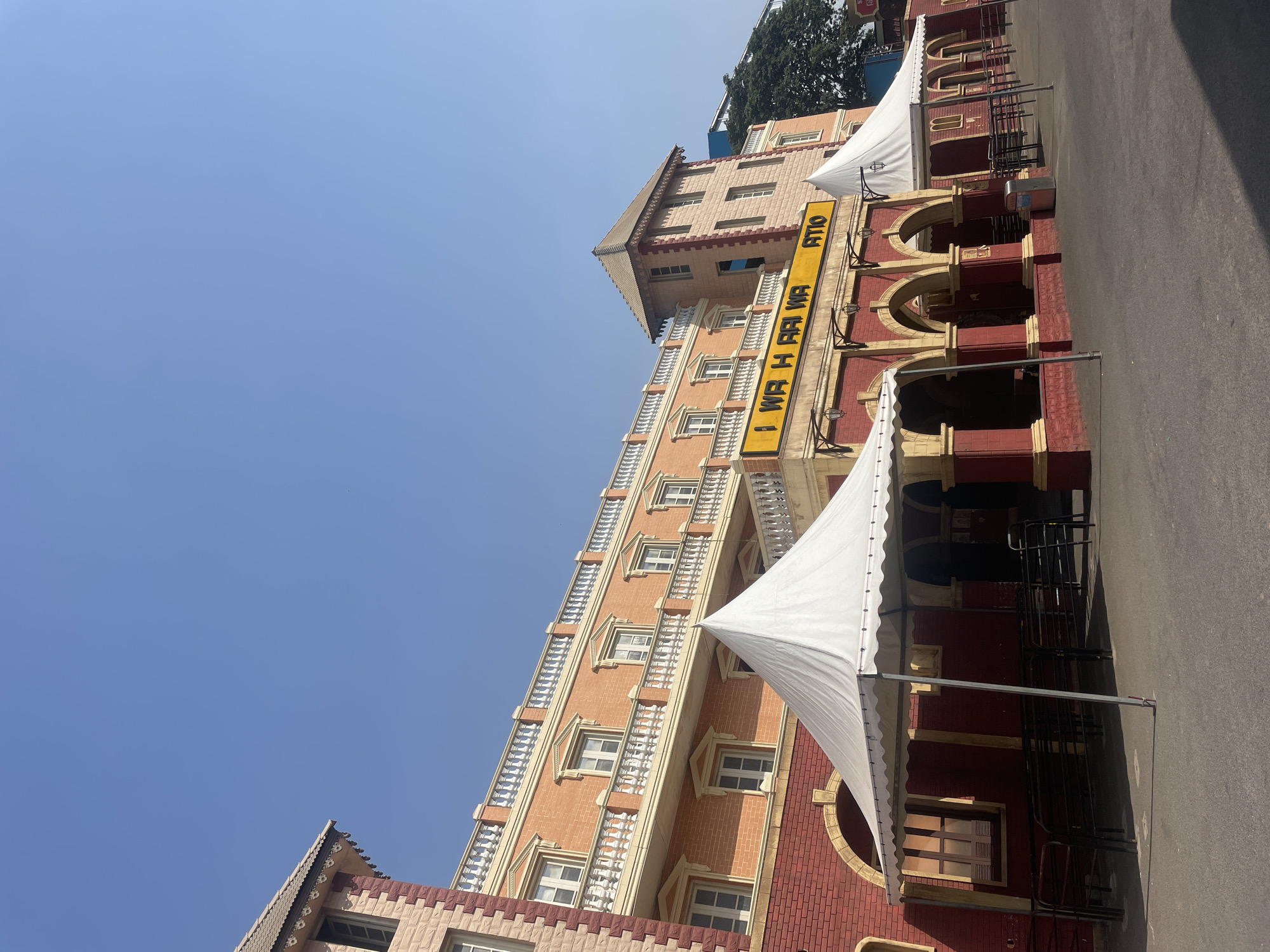 A tall, multi-story building is depicted with a combination of red brick and peach-colored textured facade. A large yellow sign vertically displays the text "I WR H RAI WA ATIO." The building features arched openings and decorative railings on some floors. White tents are positioned on the right side of the image, adjacent to the building. The ground is paved, and the sky is clear blue. Based on the signage, the location is potentially in Anajpur, India.