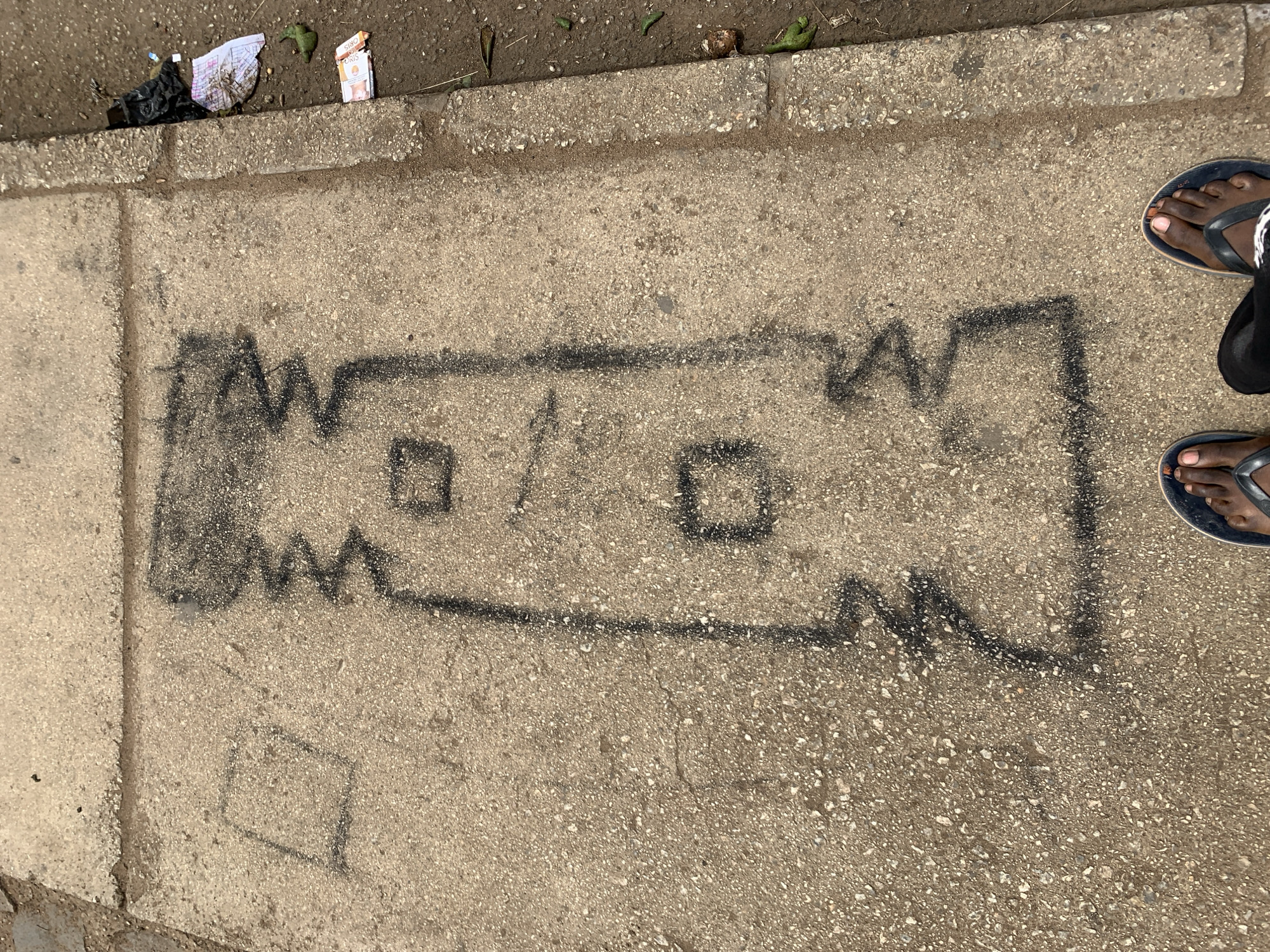 The scene is a concrete sidewalk in Rafin Par, Nigeria. There is a black graffiti drawing on the sidewalk of an object with two squared openings and jagged edges, resembling an industrial cutting tool or a saw blade. At the top edge of the frame, there is litter including paper and a possible food wrapper on the sidewalk's edge. A pair of feet wearing blue flip-flops is partially visible on the right side of the frame. The overall tone is neutral, suggesting a public space in an urban environment.