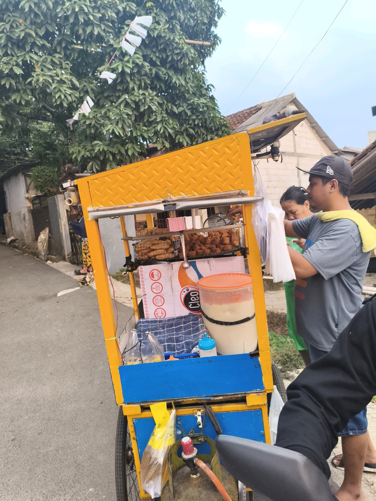 This is a photograph of a street vendor in Depok, Indonesia, operating a mobile food cart. The cart is predominantly bright yellow with blue accents and appears to be on wheels, suggesting it can be moved. On display are various food items, including what look like fried snacks arranged in wire baskets. A large, clear plastic container filled with a milky-white liquid, possibly a drink or sauce, is also visible.
Two individuals are interacting with the cart. A man wearing a grey t-shirt, a dark baseball cap, and a yellow towel draped over his shoulder is on the right side of the image, holding a white plastic bag. He appears to be serving or packaging food. Behind him, a woman with dark hair pulled back is also near the cart, looking down. The background reveals a narrow street lined with buildings, some with tiled roofs, and lush green trees. Small white flags or banners are strung across a tree branch above the cart. The overall scene depicts a typical street food vending operation in an urban or suburban Indonesian setting.