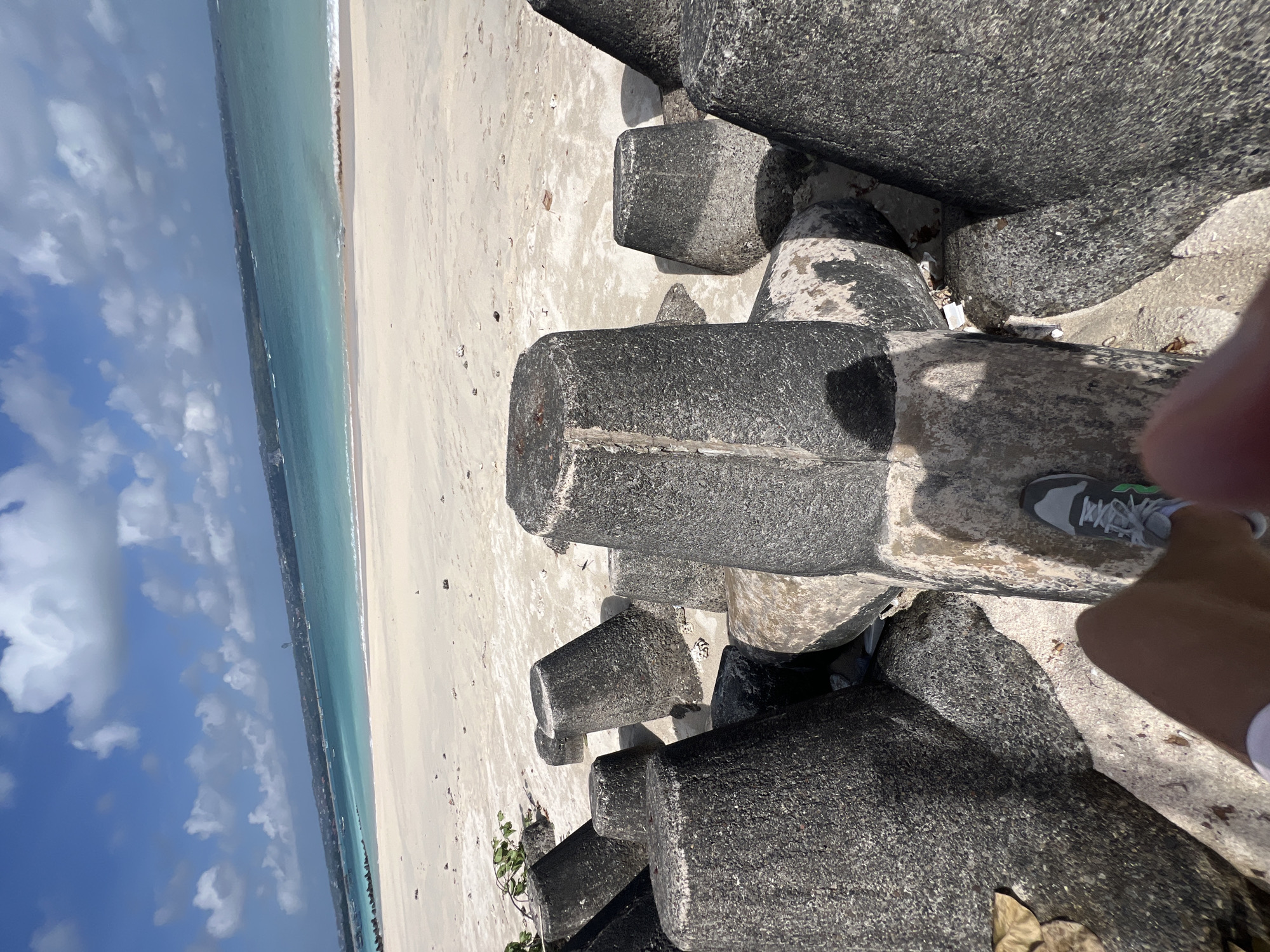 A first-person perspective shot of a person standing on a beach in Kuta, Bali, Indonesia. The image captures a segment of a beach with a cluster of concrete tetrapods in the foreground, used as a breakwater. The tetrapods are dark gray and weathered, some showing signs of algae and erosion. Beyond the tetrapods, a sandy beach stretches towards the turquoise ocean. The water is calm with gentle waves lapping the shore. In the distance, a hazy coastline is visible with buildings and vegetation. The sky is a bright blue with scattered white clouds. The lower part of the image shows the person's legs and feet, wearing athletic shoes and shorts, standing on a tetrapod. The overall scene depicts a sunny day at a tropical beach.