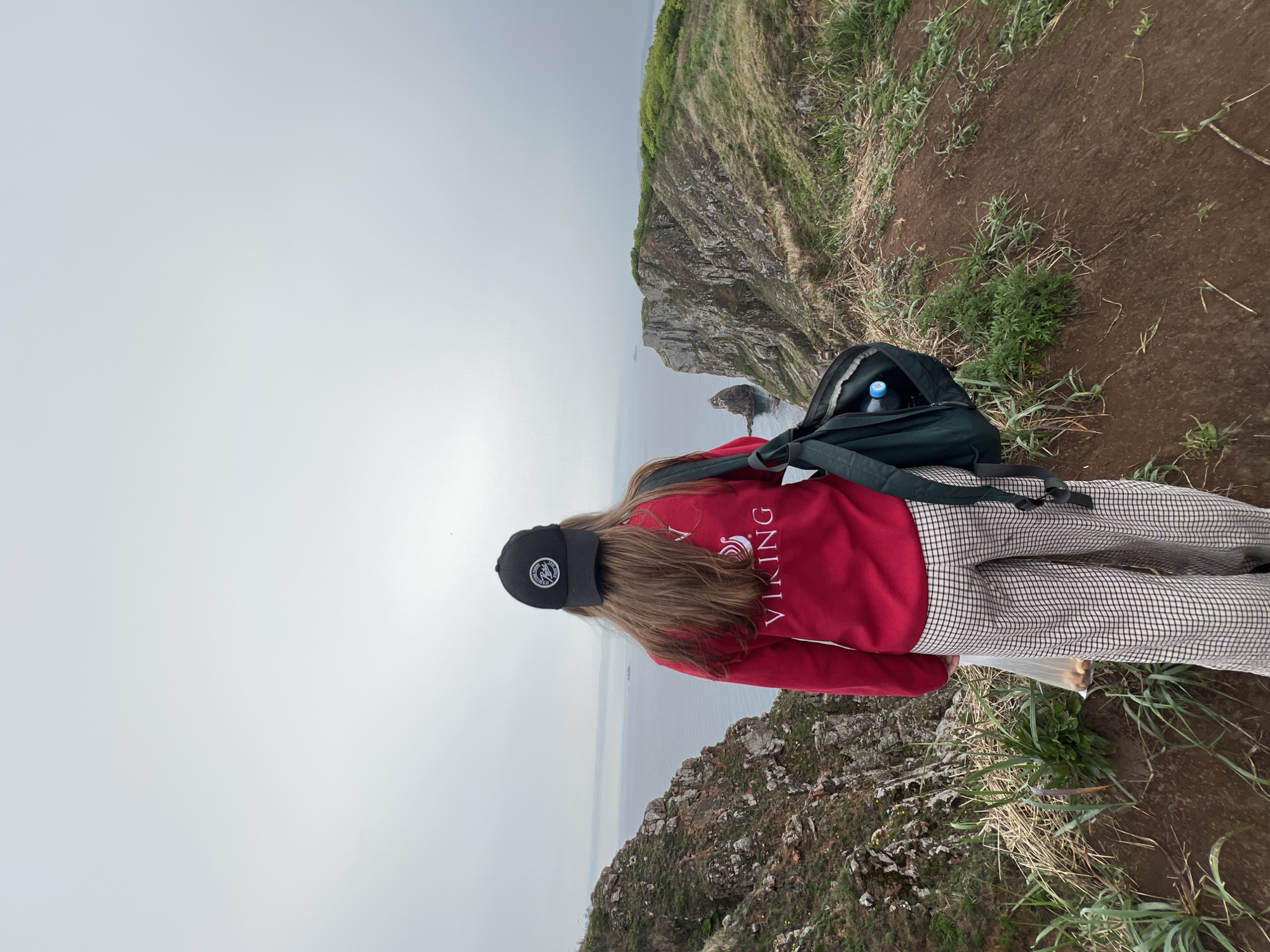 The image depicts a person standing on a cliff overlooking the sea in Russkiy Island, Russia. The person wears a black cap, a red sweater with the word "VIKING" on the back, and plaid pants. A backpack rests on their back. The landscape features rocky cliffs, grassy terrain, and a foggy or overcast sky. The sea is visible below, with some rocky formations emerging from the water.