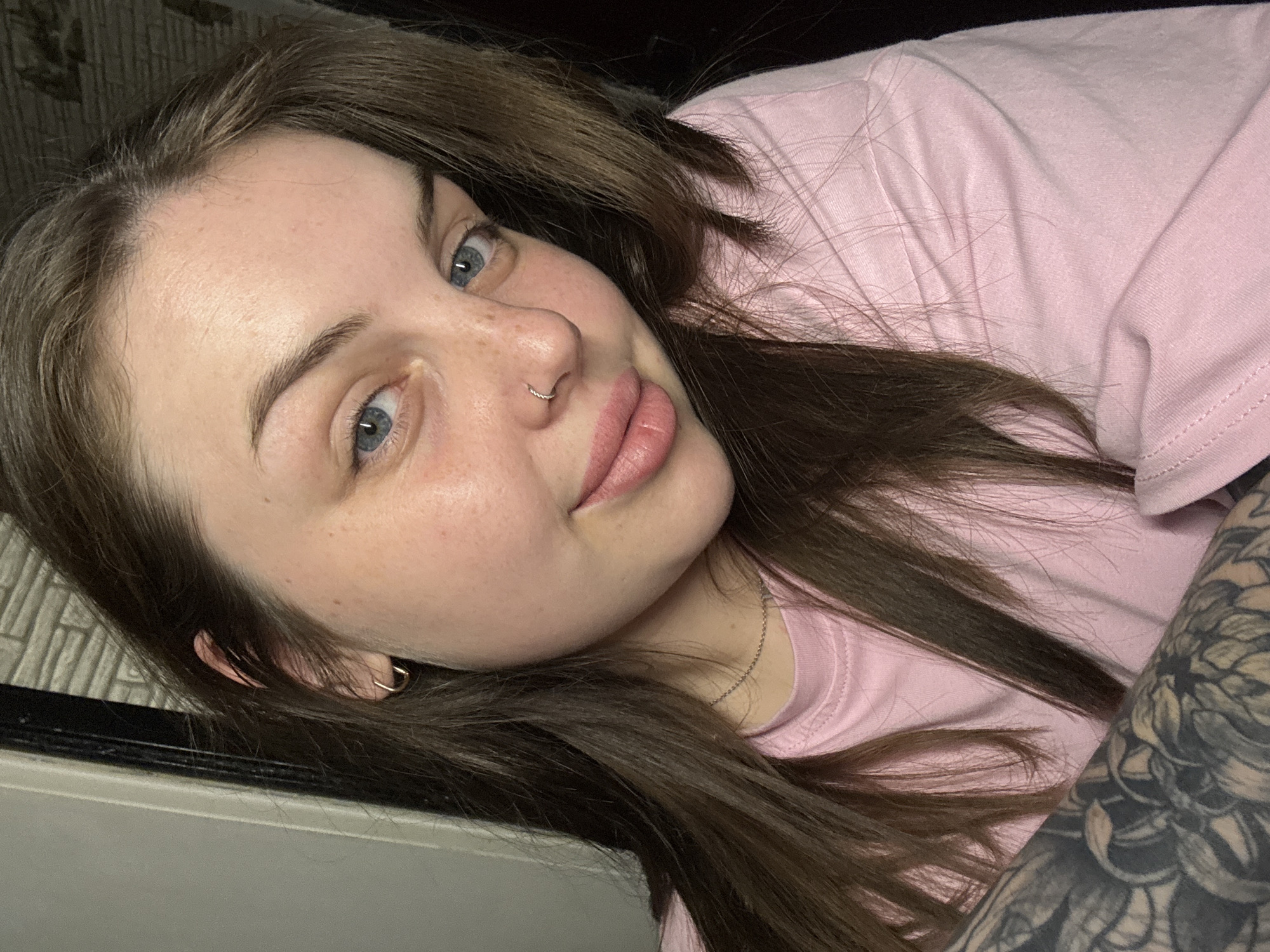 A woman lies on a surface with her head resting against a gray and black tile-like surface. She wears a light pink t-shirt. Her hair is brown and long, spread around her head and shoulders. She has blue eyes, fair skin with freckles, and a silver nose ring. A silver hoop earring is visible in her left ear. Her arm, covered in a dark tattoo, is also visible. The background behind her is dark. The location is unknown.