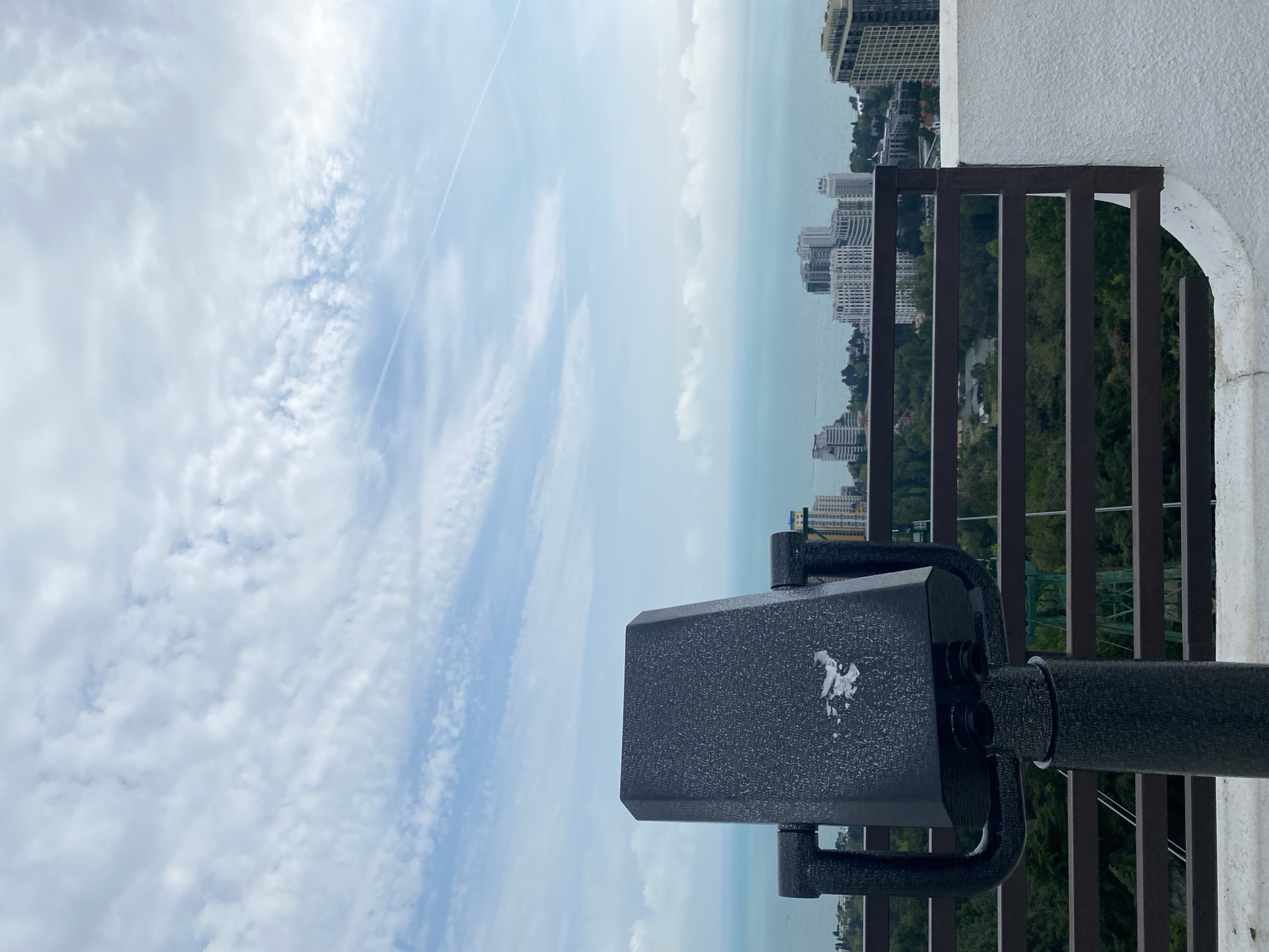 From an elevated point of view, the image presents a cityscape scene, possibly in Sochi, Russia, based on the architectural style. The viewpoint is from a balcony featuring a black, coin-operated viewing binoculars pointed towards the skyline. A brown metal railing divides the foreground from the vista, which includes high-rise buildings, interspersed with green vegetation. The skyline fades into a light blue horizon, merging with the sky dotted with white clouds.