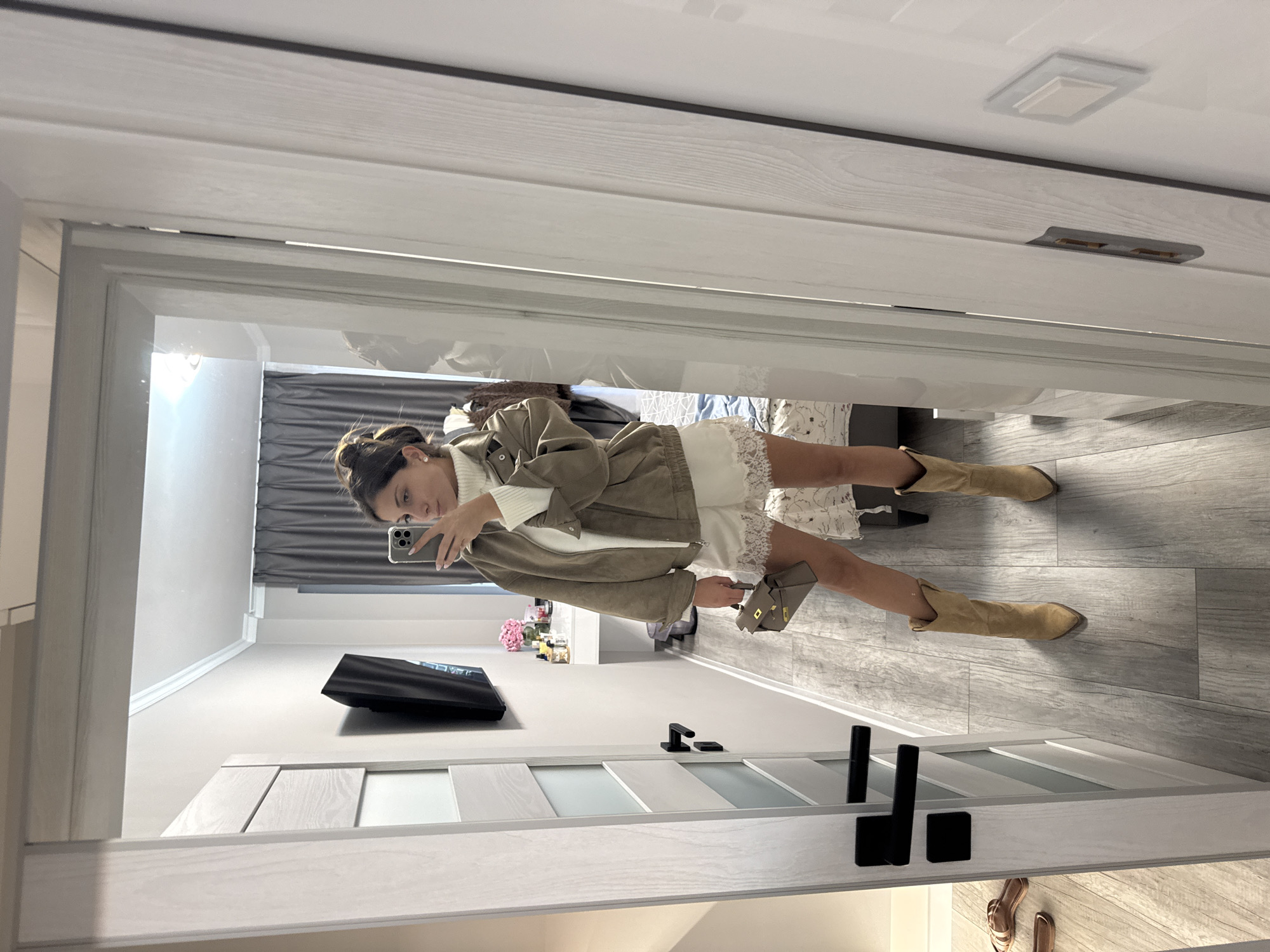 A young woman is taking a mirror selfie in what appears to be a bedroom in Burgas, Bulgaria. She is wearing a white turtleneck sweater, a light brown bomber jacket, white lace shorts, and tan cowboy boots. She is holding a small gray handbag. The room features a large mirror, a white dresser with decorative items, a bed, and gray curtains. The floor is wood-paneled, and the walls are light gray. A black-handled door is visible to the left, and a white door with wooden accents is to the right.