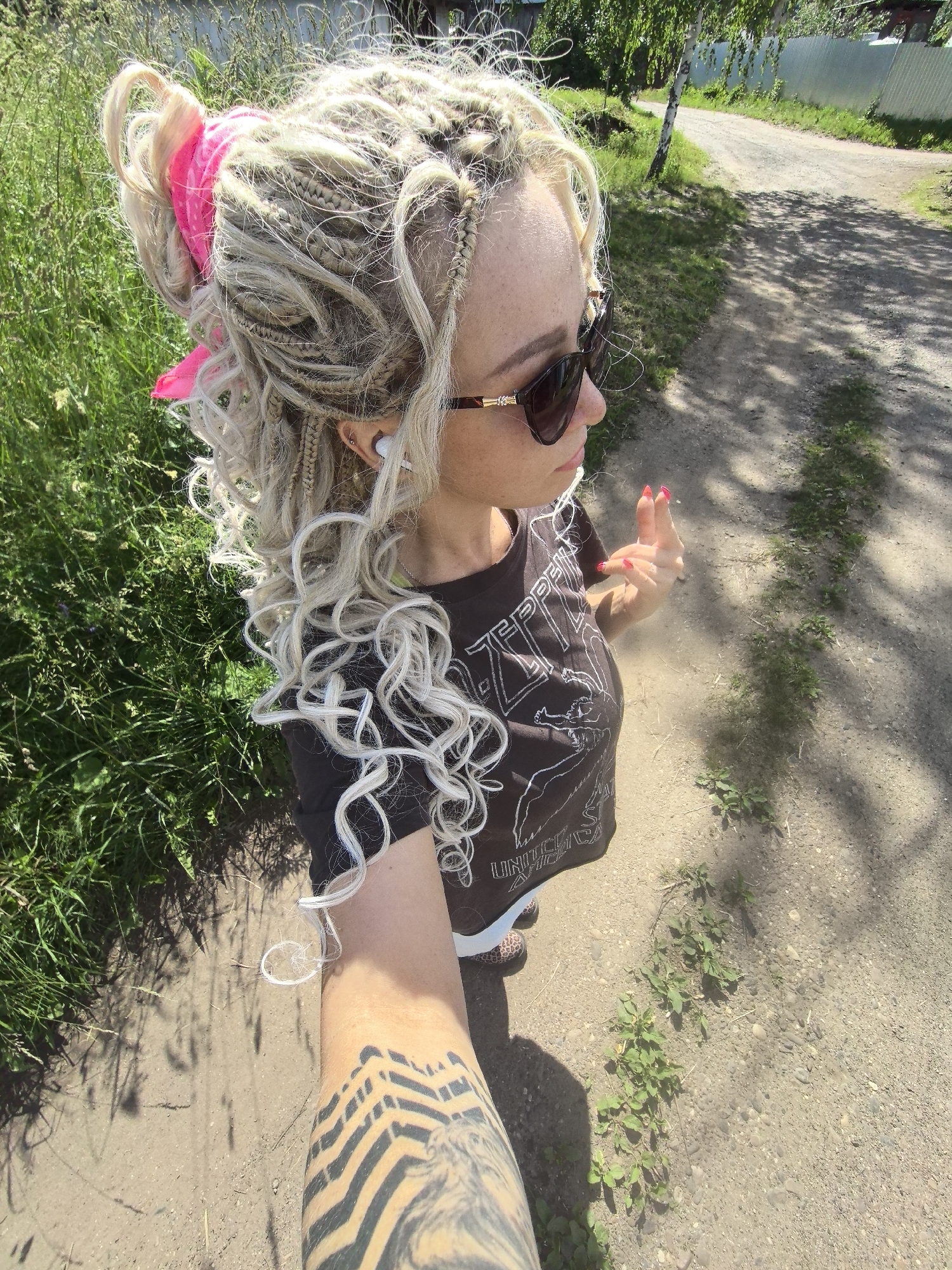 The image depicts a woman standing outdoors on a dirt path. She has blonde, braided hair styled into a high bun with a pink bandana. She is wearing a dark t-shirt with a graphic print, sunglasses, and appears to have earbuds in. Her left arm, prominently visible in the foreground, is covered in a large black tattoo. She is holding her right hand up, showing manicured nails. The background shows green grass, trees, and a fence. The path appears to be in a rural or suburban area, possibly on the outskirts of Noviy, Russia, given the visual context of the surroundings which are consistent with Russian countryside. The lighting suggests it is daytime and sunny.