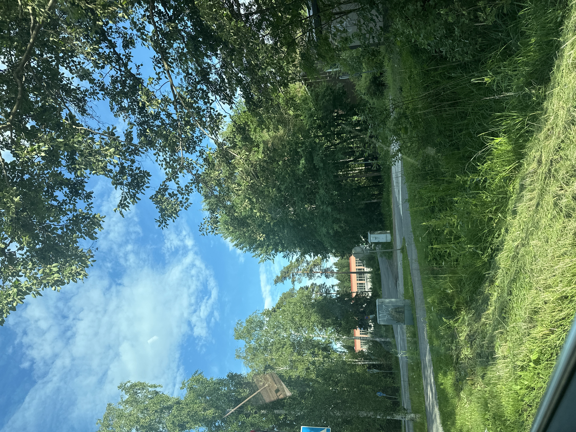 The image shows a view of a street with trees and a building in the background. The sky is blue with scattered white clouds. In the foreground, there is a grassy area with some cut grass. A road runs through the scene, with a building visible on the other side. There are also some utility boxes and signs along the road. The overall impression is a suburban or semi-rural setting on a sunny day.