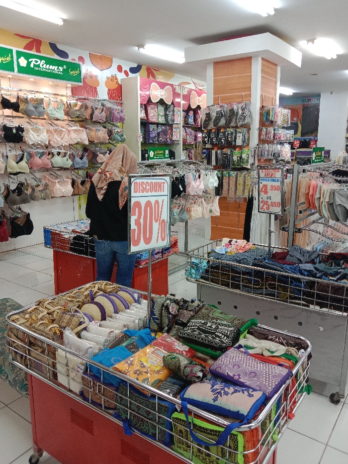 The scene depicts the interior of a brightly lit retail store located in Langenharjo, Indonesia. One individual is present, seen from the back, wearing a light brown patterned hijab, a black long-sleeved shirt, and blue jeans. They are standing near a low red display table filled with packaged items.
The store is arranged with various merchandise. The left wall displays multiple rows of bras in assorted colors (black, beige, pink, light blue, white) on hangers, beneath signage for "Plums International." A prominent white sign with red lettering reading "DISCOUNT 30%" stands on a pole in the mid-ground. To the right of the individual, additional clothing items, including packaged socks or hosiery and various underwear, are displayed on wall-mounted and freestanding racks. Pricing signs, displaying "4.950" and "25.450" in Indonesian Rupiah, are also visible.
In the foreground, two large, rectangular metal-framed bins on wheeled bases are present. The left bin, on a red base, is filled with small woven bags, packaged goods, and folded textiles, possibly including small prayer mats. The right bin, on a grey base, contains a assortment of folded clothing or textile items in various colors and patterns. The store features white walls, with a central column accented with light orange wood-grain paneling, and a light-colored tiled floor.
