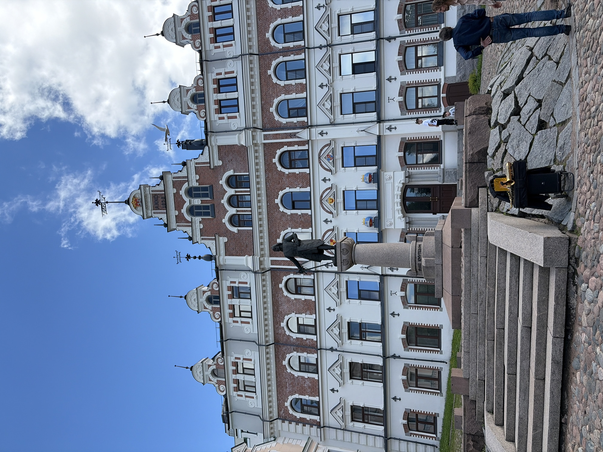 An outdoor daytime scene captures a multi-story building with ornate brick and white facade, featuring numerous arched windows. A stone pedestal stands in front of the building, topped with a bronze statue of a man holding a staff. To the left of the pedestal, a set of stone stairs leads up to a paved area. In the foreground, a cobblestone ground is visible, with a cluster of large rocks on the right. On top of these rocks, a musical instrument, possibly a saxophone, is set up on a stand next to a black speaker. Four people are present in the scene: two individuals stand near the rocks, looking towards the building and the musician; one person, a child, is seated on a rock near the stairs; and another person, a man wearing a dark blue shirt and jeans, stands near the rocks, facing away from the camera. The sky above is blue with scattered white clouds.