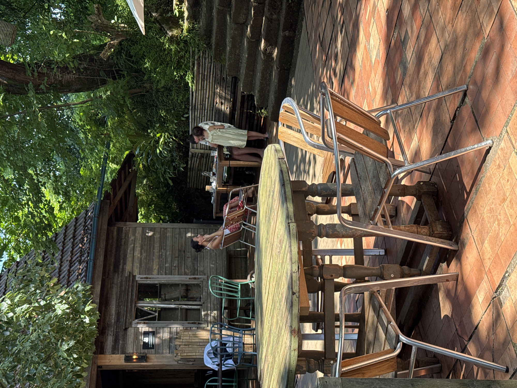 In Buknari, Georgia, an outdoor seating area of a restaurant features a weathered wooden building with a tiled roof. In the foreground, a large, round, distressed green wooden table is surrounded by several chairs with metal frames and wooden seats. The chairs are positioned on a red tiled patio. In the background, a woman with dark hair tied back sits in a chair near the building, looking to the side. Further back, a man stands with a young child wearing a striped dress, interacting at another table. A set of stone steps is visible to the right, leading upwards into a lush, green, forested environment. The scene is bathed in dappled sunlight, indicating a daytime setting.