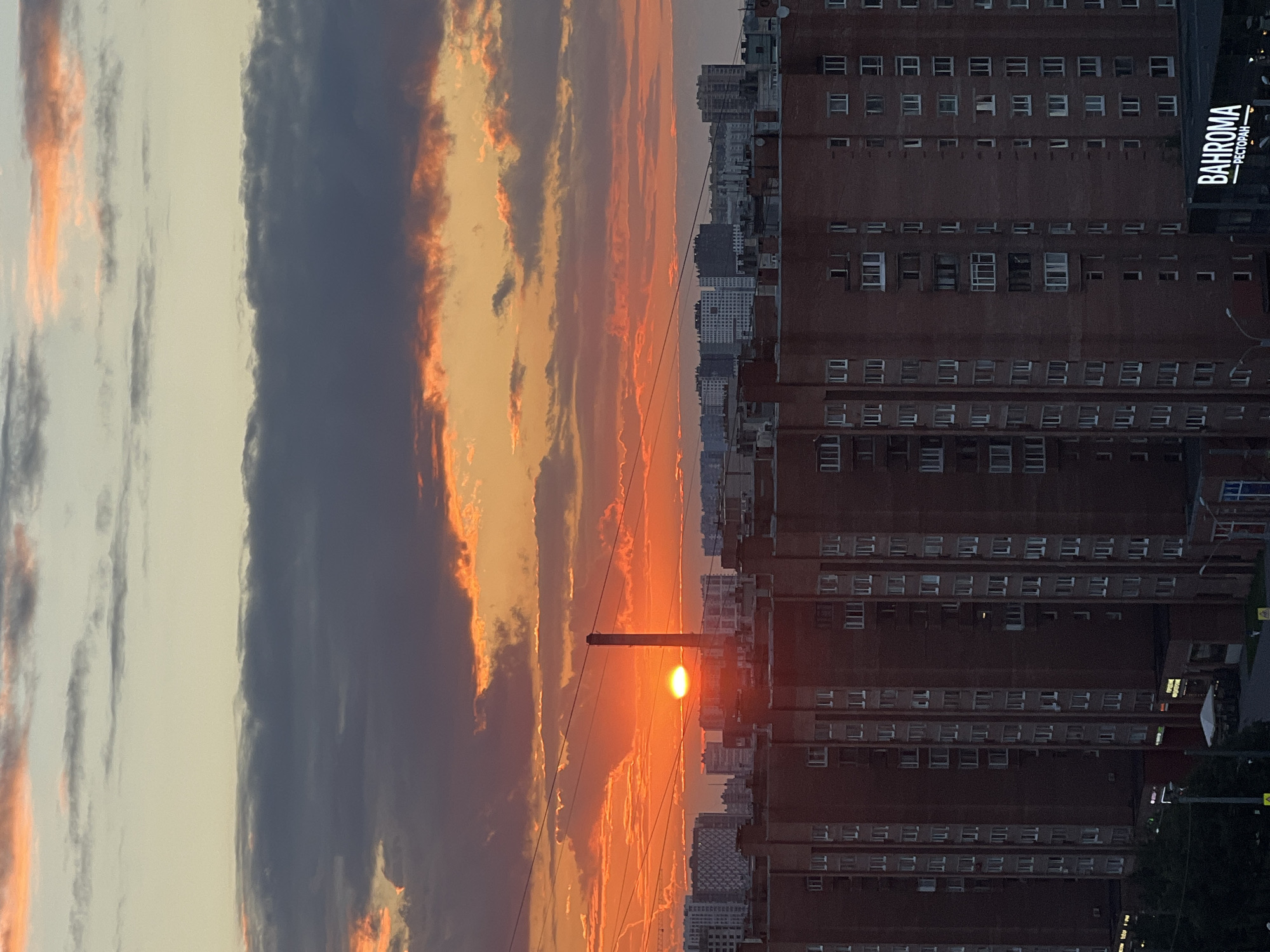 The image depicts a cityscape at sunset. The sky is filled with vibrant orange and pink clouds, illuminated by the setting sun, which appears as a bright, small orb. A silhouetted utility pole stands in the foreground, with wires stretching across the frame. To the right of the frame, a large, brick apartment building dominates the scene. The building has numerous windows, many of which are illuminated, suggesting it is inhabited. Another, taller building is visible in the distance, also with many windows. The overall mood is serene and captures the beauty of a sunset over an urban environment. Visible text on the brick building reads "BAHROMA РЕСТОРАН", indicating a restaurant named Bahroma. The time of day is clearly sunset, and the weather appears clear, with dramatic cloud formations. The location is urban, with evident residential and commercial structures.